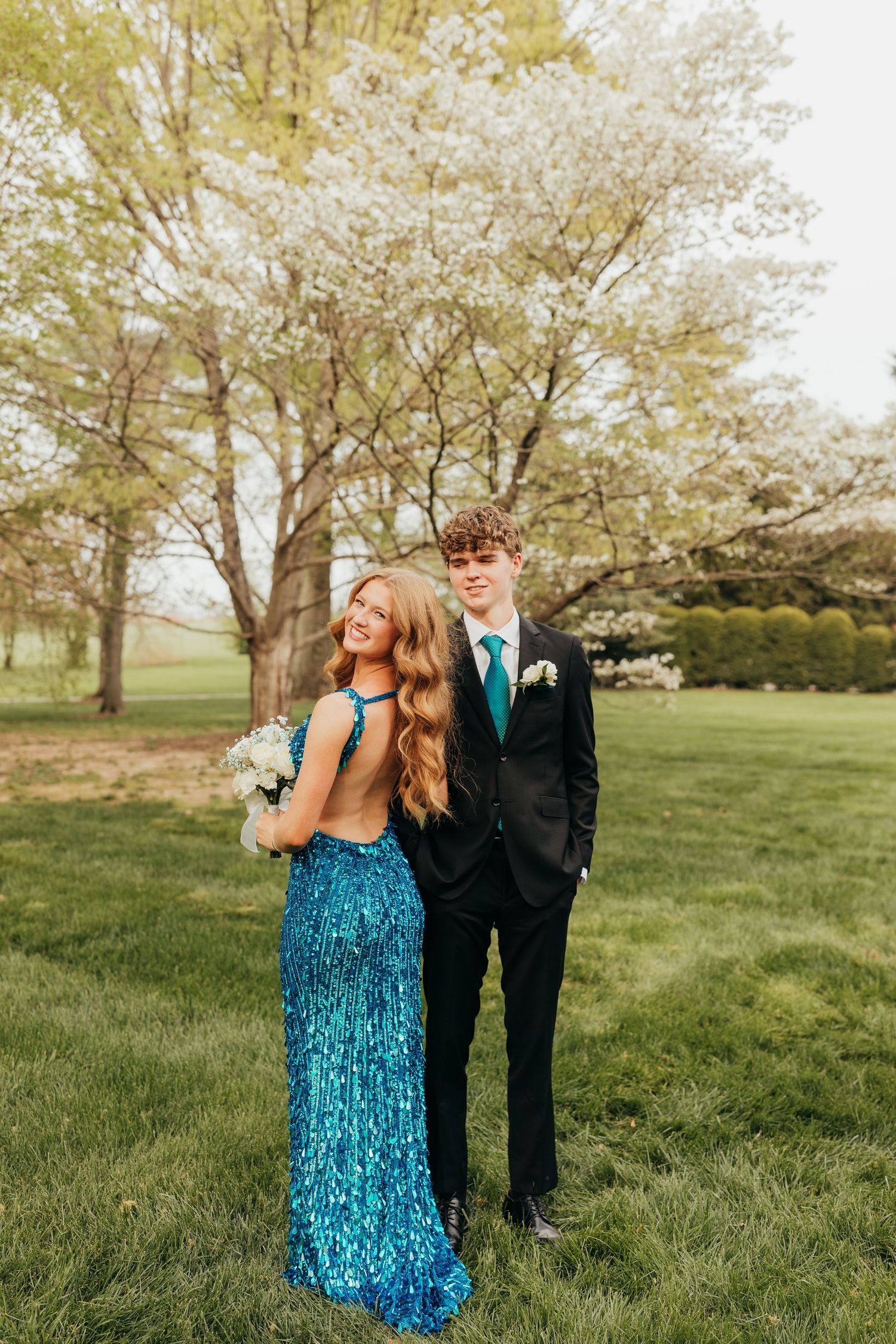 Prom couple posing outdoors, girl in backless blue sequin gown holding bouquet near blooming trees.