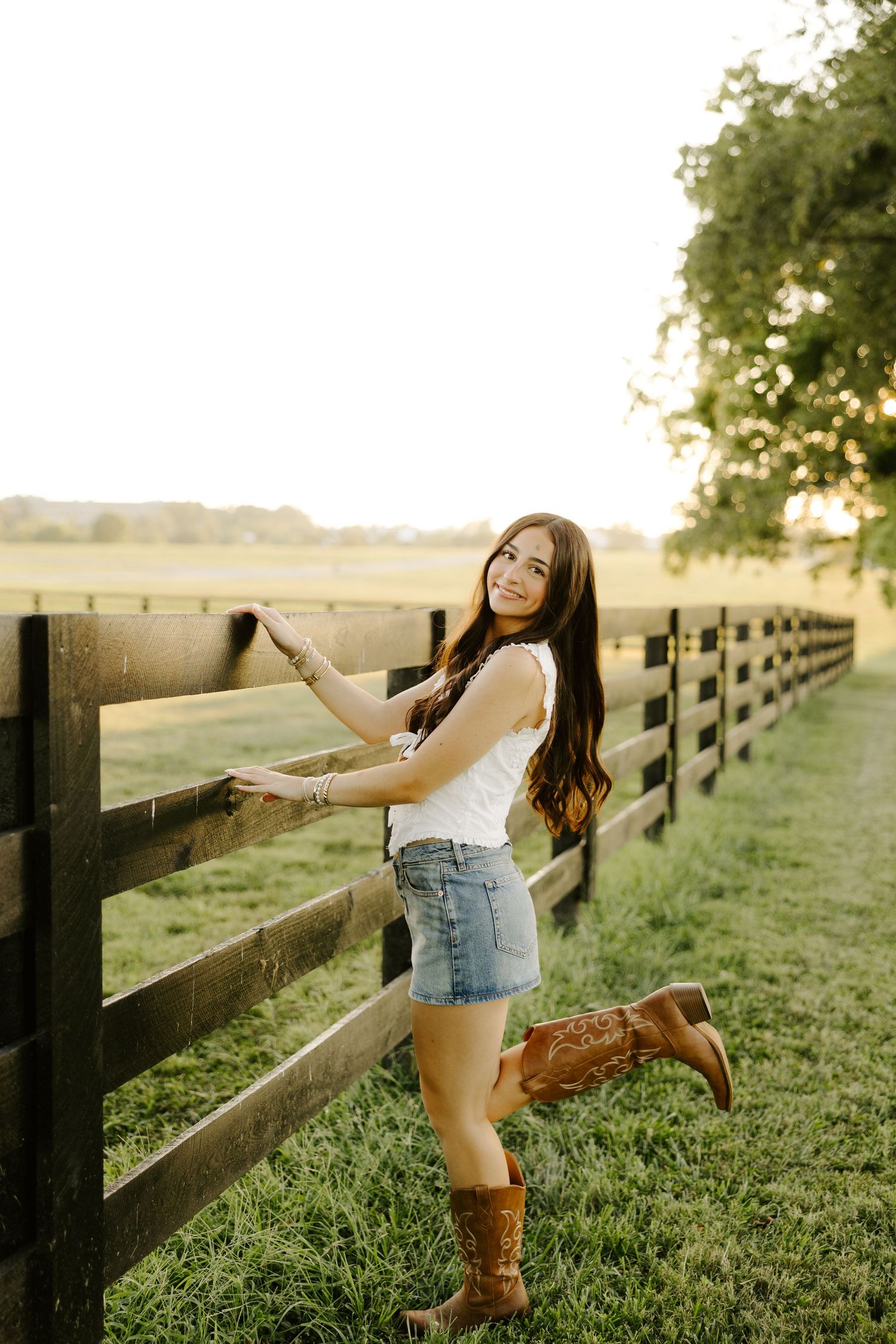 Teen girl in white top, denim skirt, and cowboy boots posing by dark wooden farm fence.