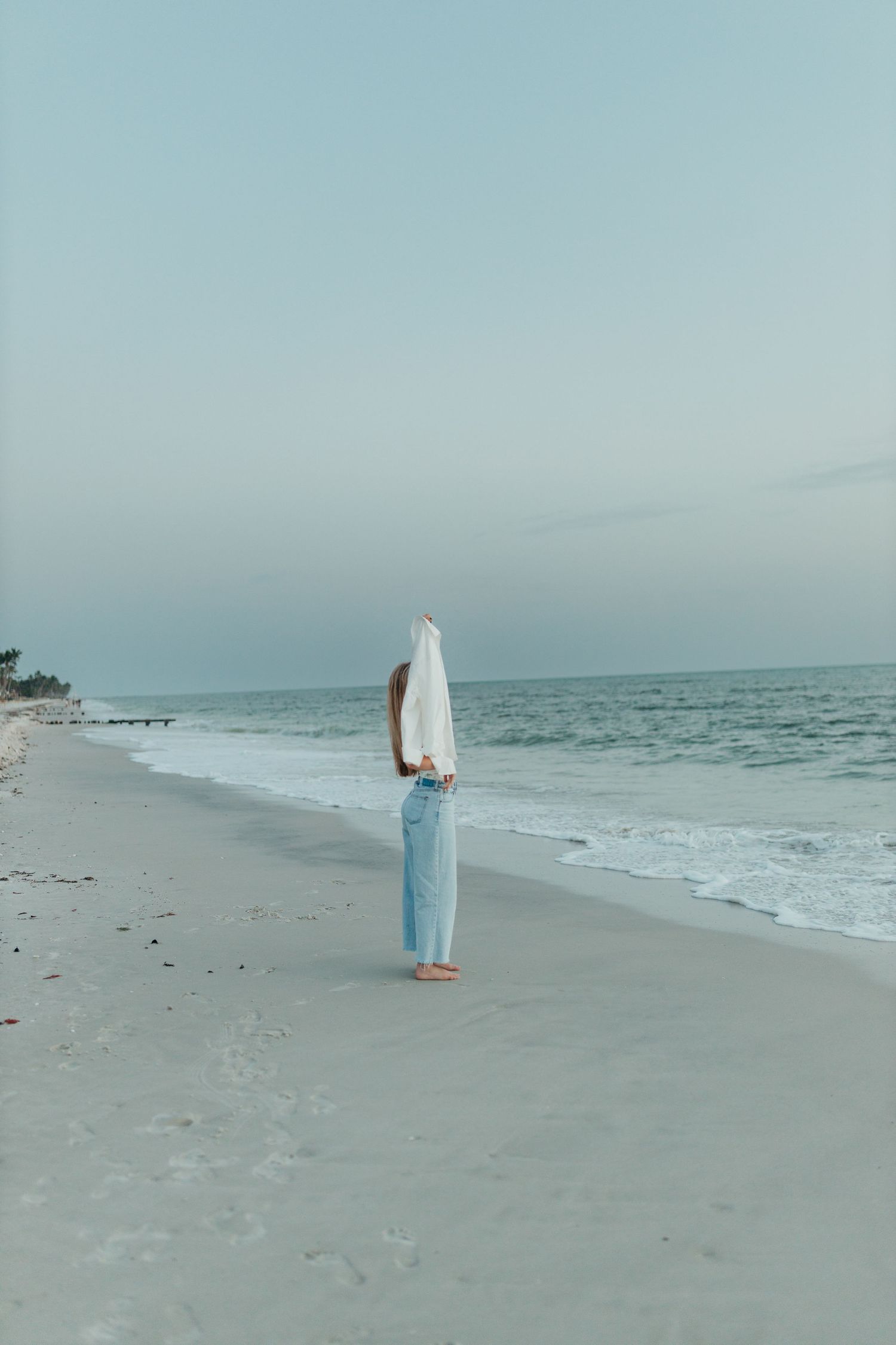 Person standing on a serene beach at dusk, lifting their white shirt above their head in blue jeans.
