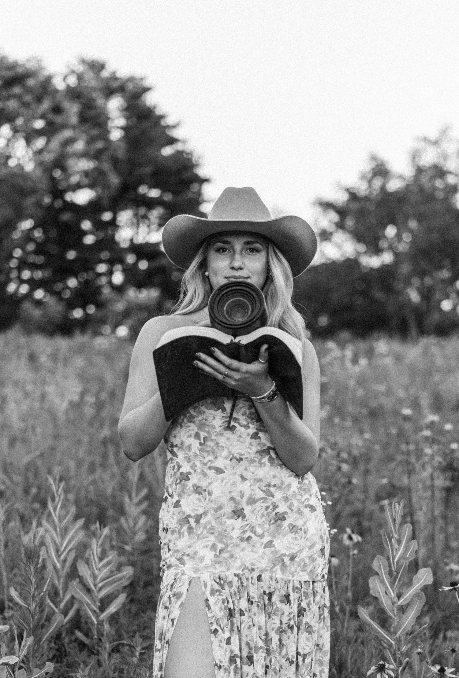 Black and white portrait of blonde woman in cowboy hat holding a camera in a field.