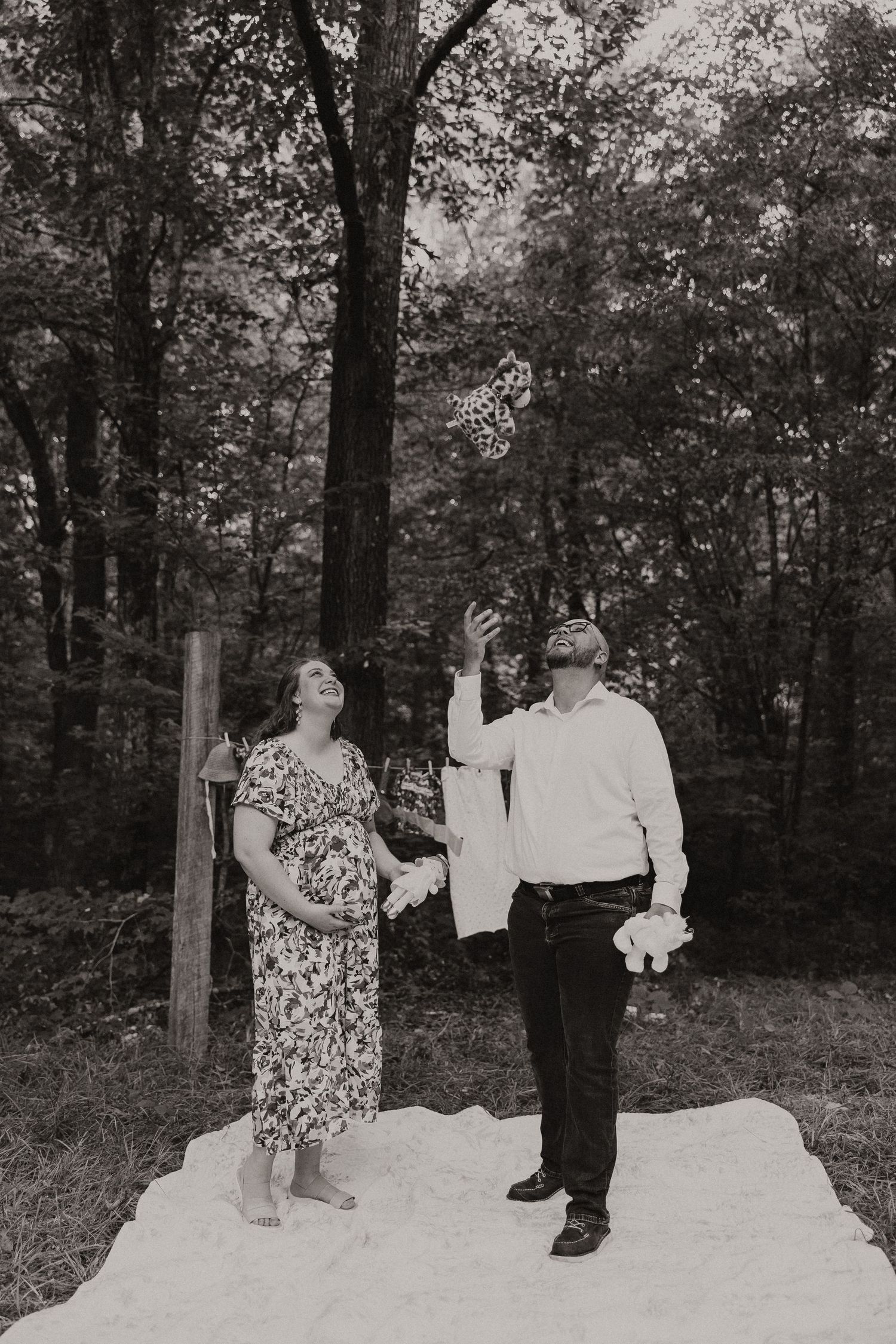 Black and white photo of pregnant couple tossing baby clothes in the air outdoors in a forest setting.