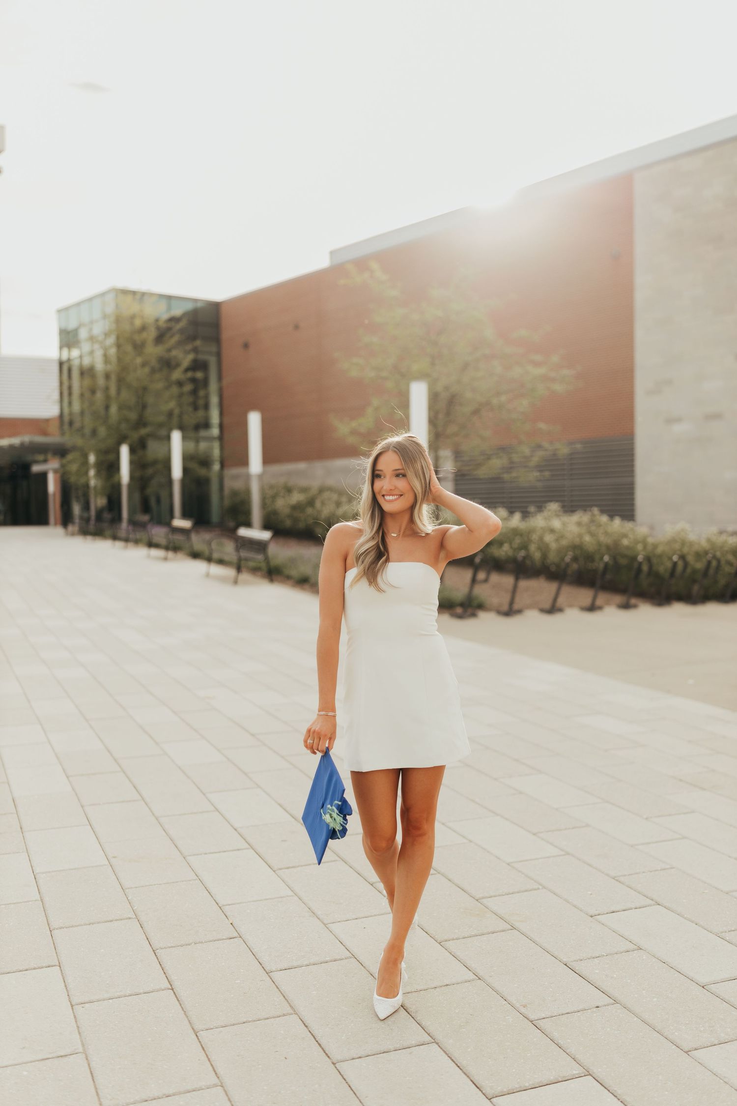 Graduate in short white dress walking on outdoor plaza holding blue graduation cap.