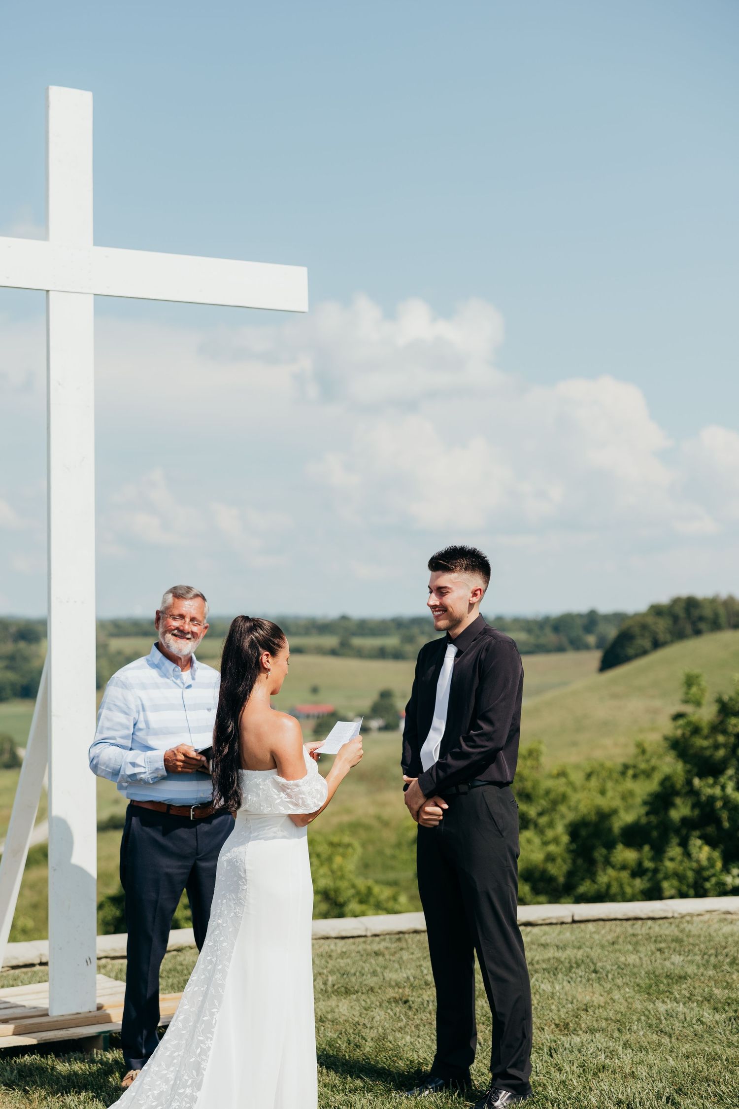 Bride reading vows to groom at outdoor ceremony beside white cross with scenic countryside backdrop.