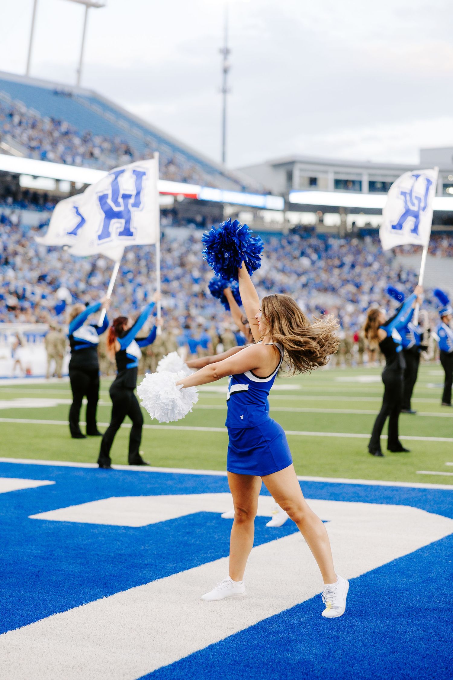 A UK cheerleader dances with blue and white pom-poms on the field during a Kentucky Wildcats football game.