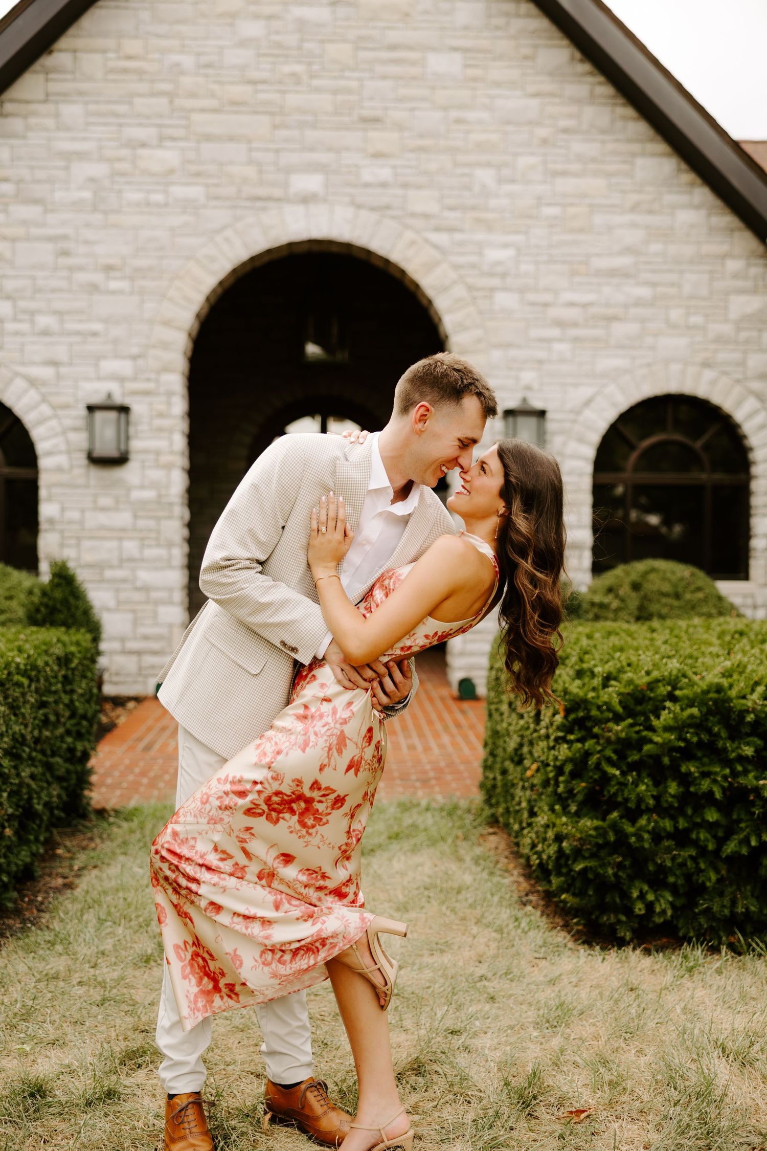 Couple sharing a romantic dip kiss in front of a stone chapel surrounded by lush green hedges.