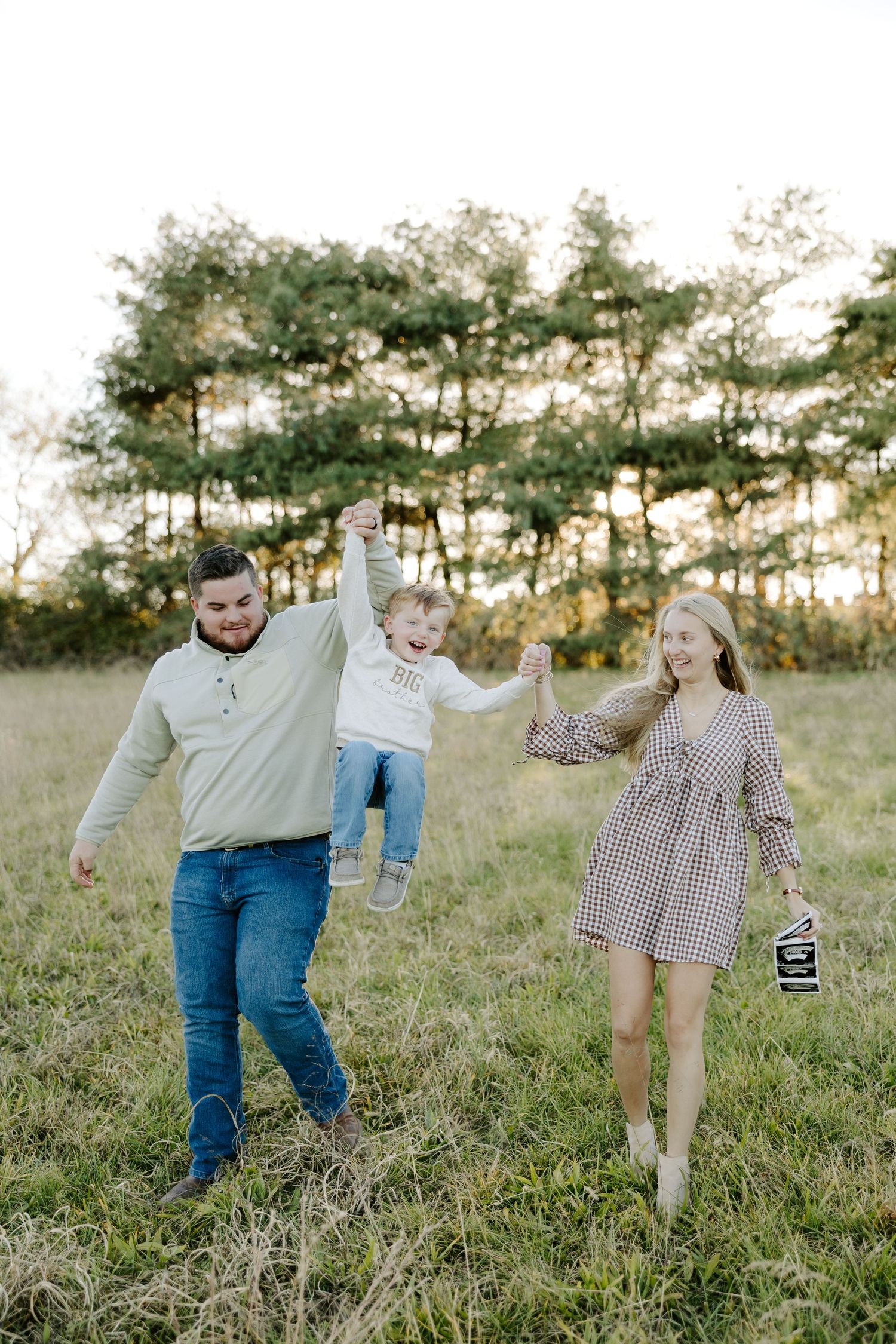 Family of three walking through sunny meadow, parents swinging laughing toddler between them holding ultrasound photos.