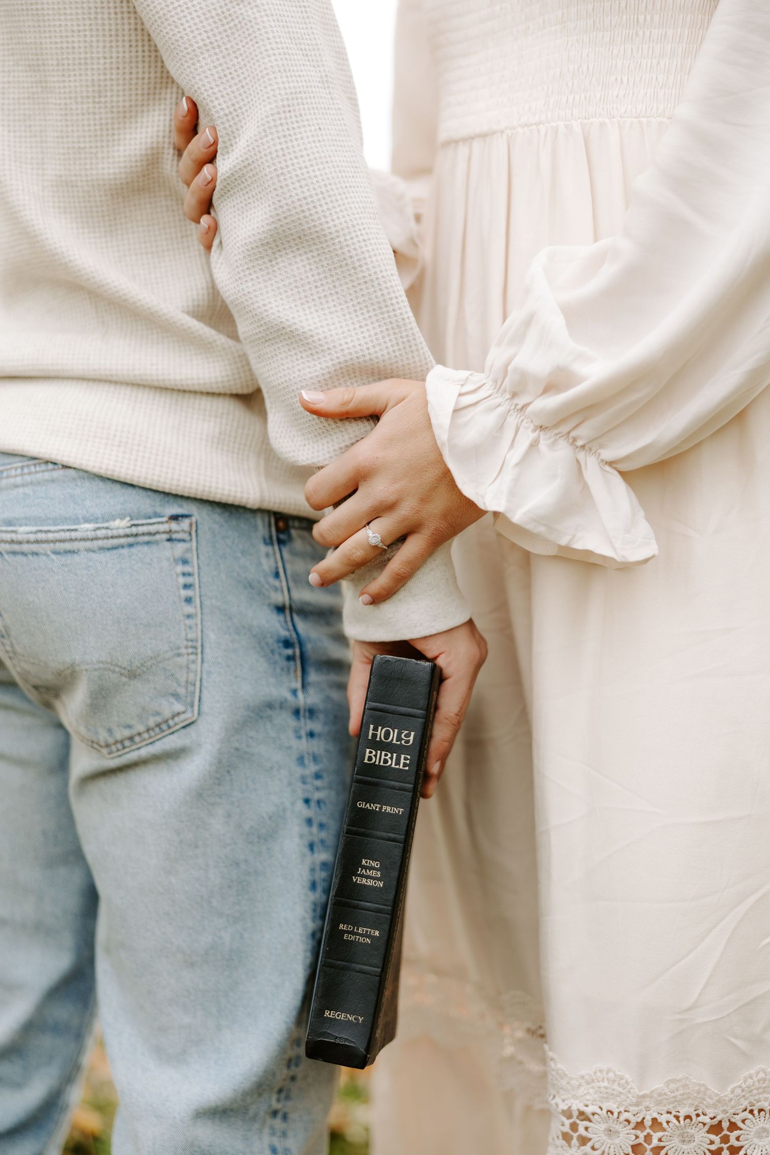 A couple stands together outdoors holding a Holy Bible, with the woman wearing an engagement ring.