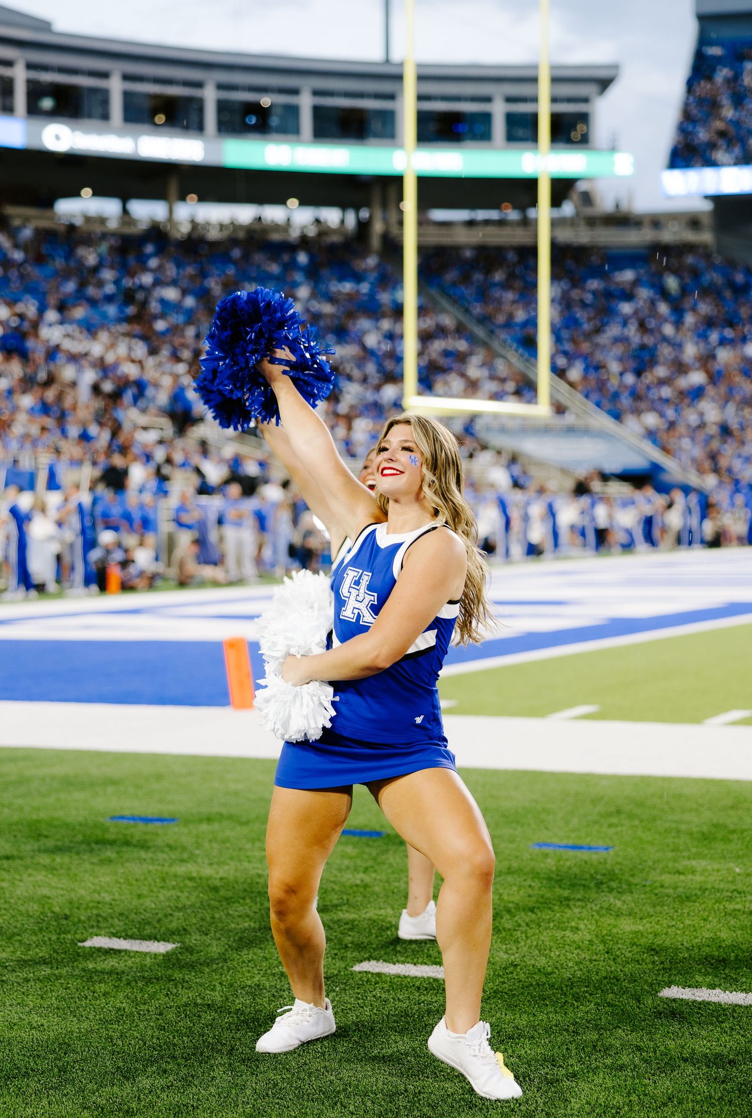 A University of Kentucky cheerleader raises blue pom-poms near the end zone during a packed Wildcats home game.