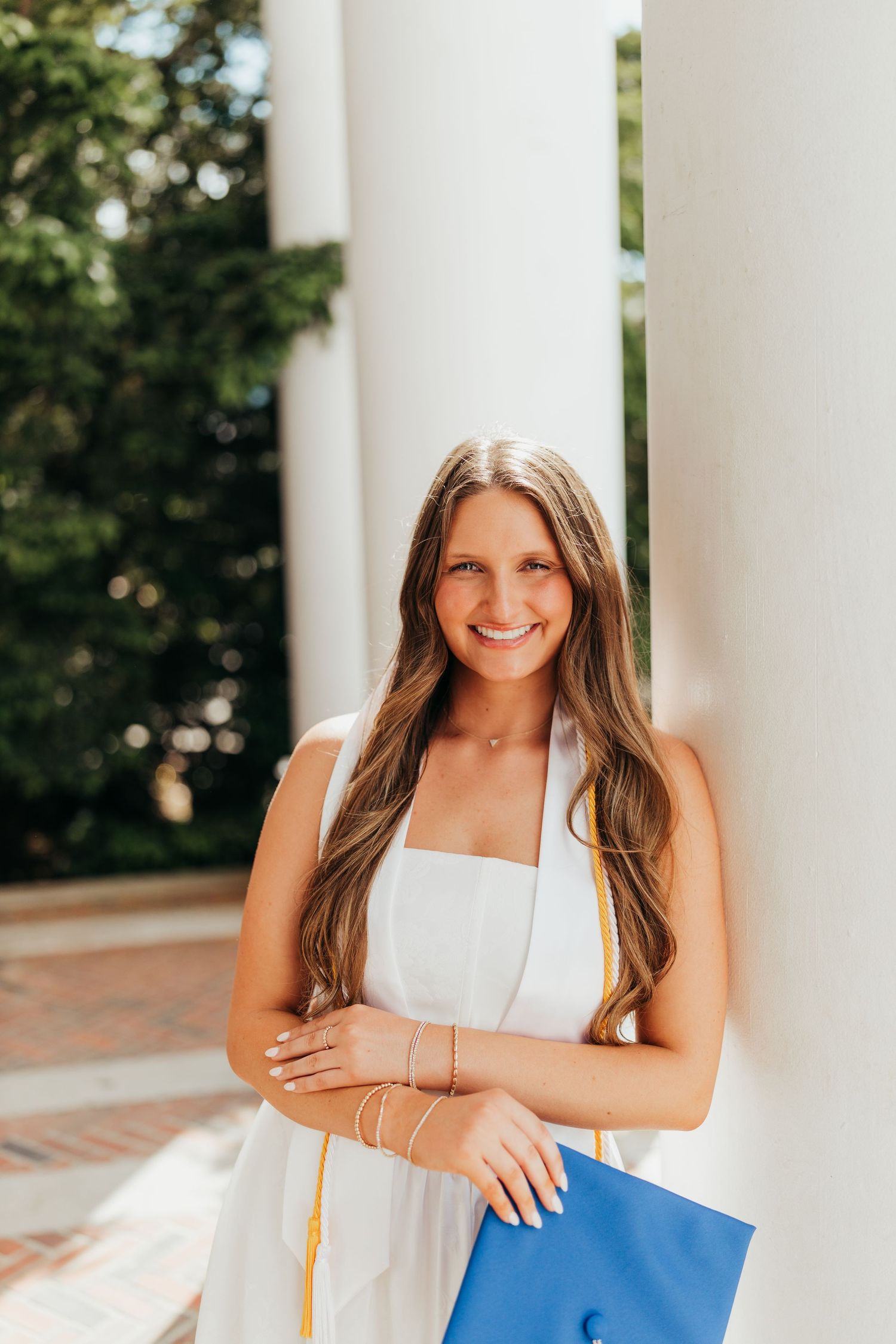 Graduate in white dress poses between classical white columns on campus.