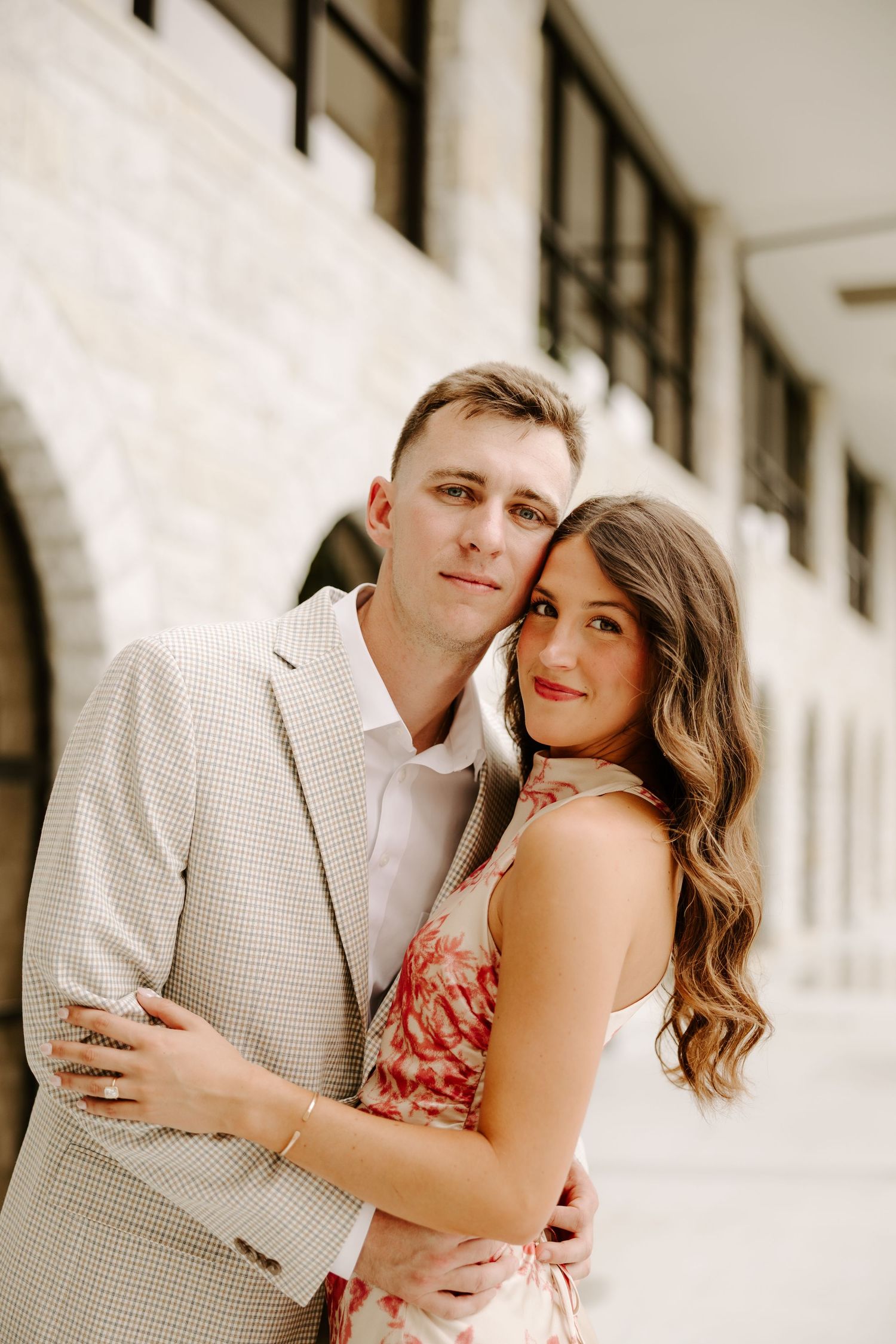 Elegant couple posing closely together under stone arches, woman in floral dress and man in beige suit.