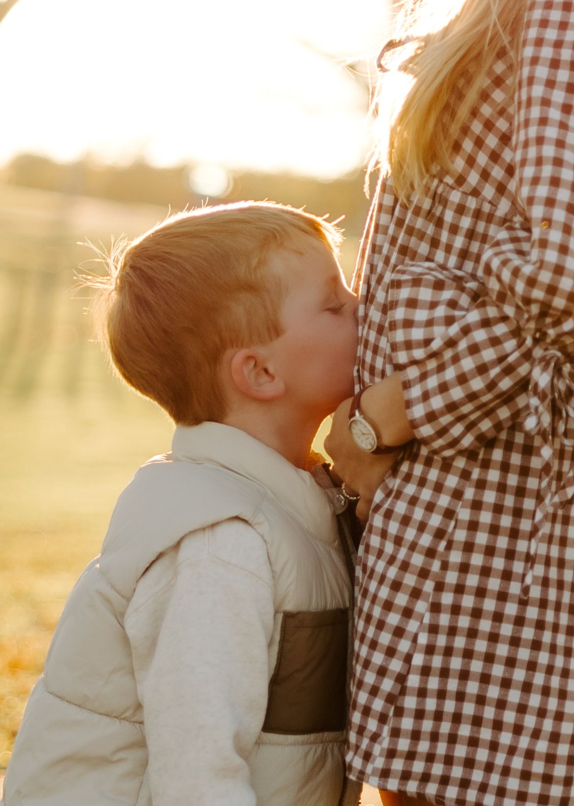 Sweet toddler kissing mother's pregnant belly in warm golden sunset light during outdoor maternity session.