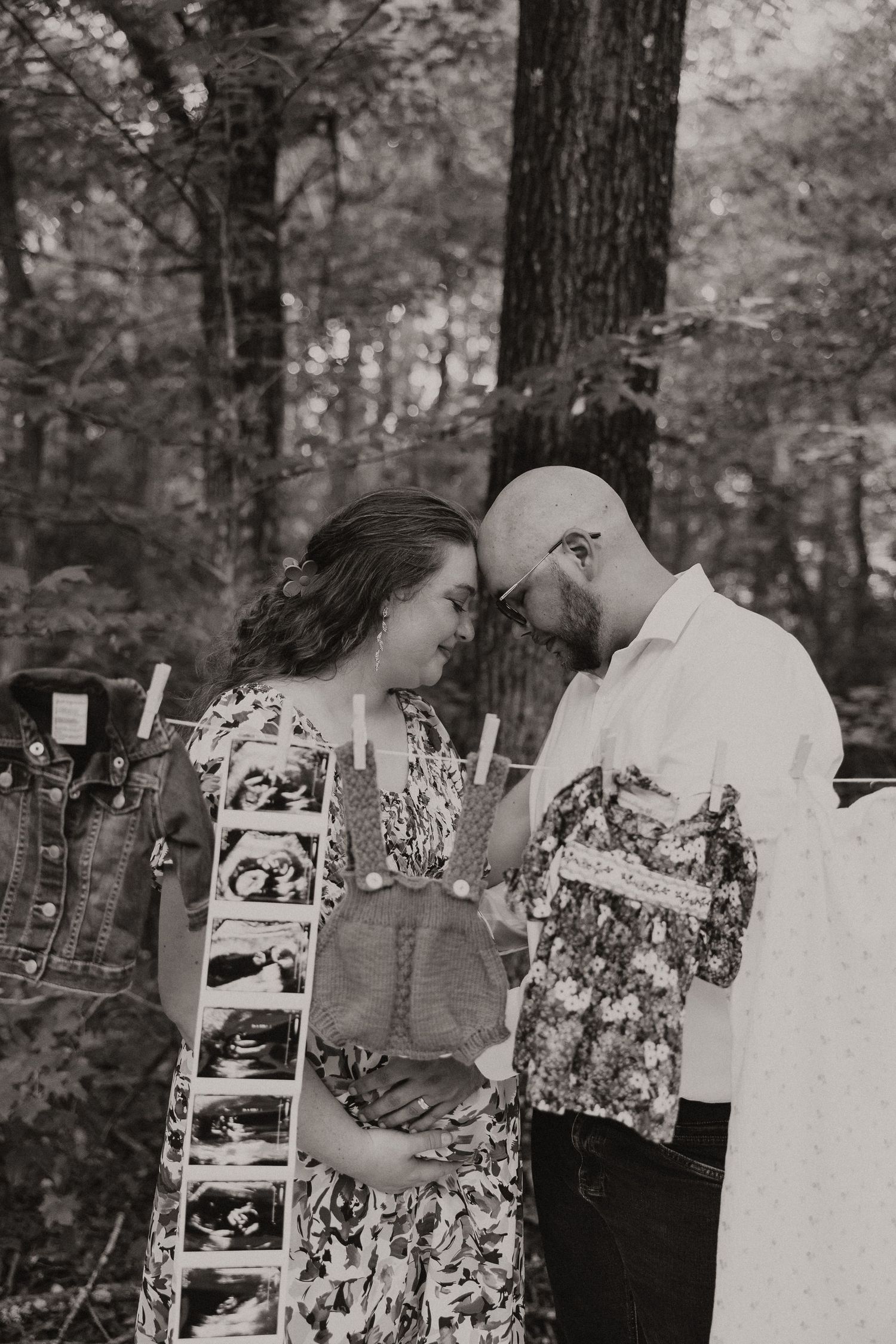 Black and white intimate maternity photo of couple touching foreheads near ultrasound photos in forest.