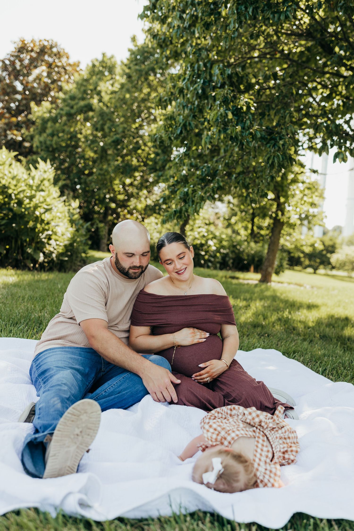 Pregnant couple sitting on blanket in park watching toddler daughter lying in front of them.