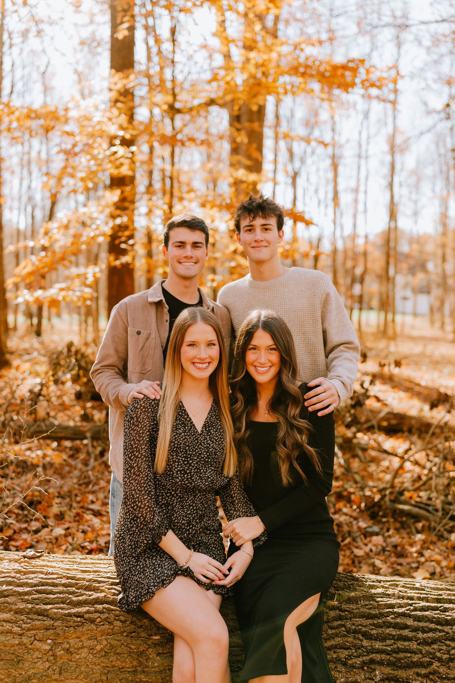 Two young men standing behind two young women posing on a log in a golden autumn forest.