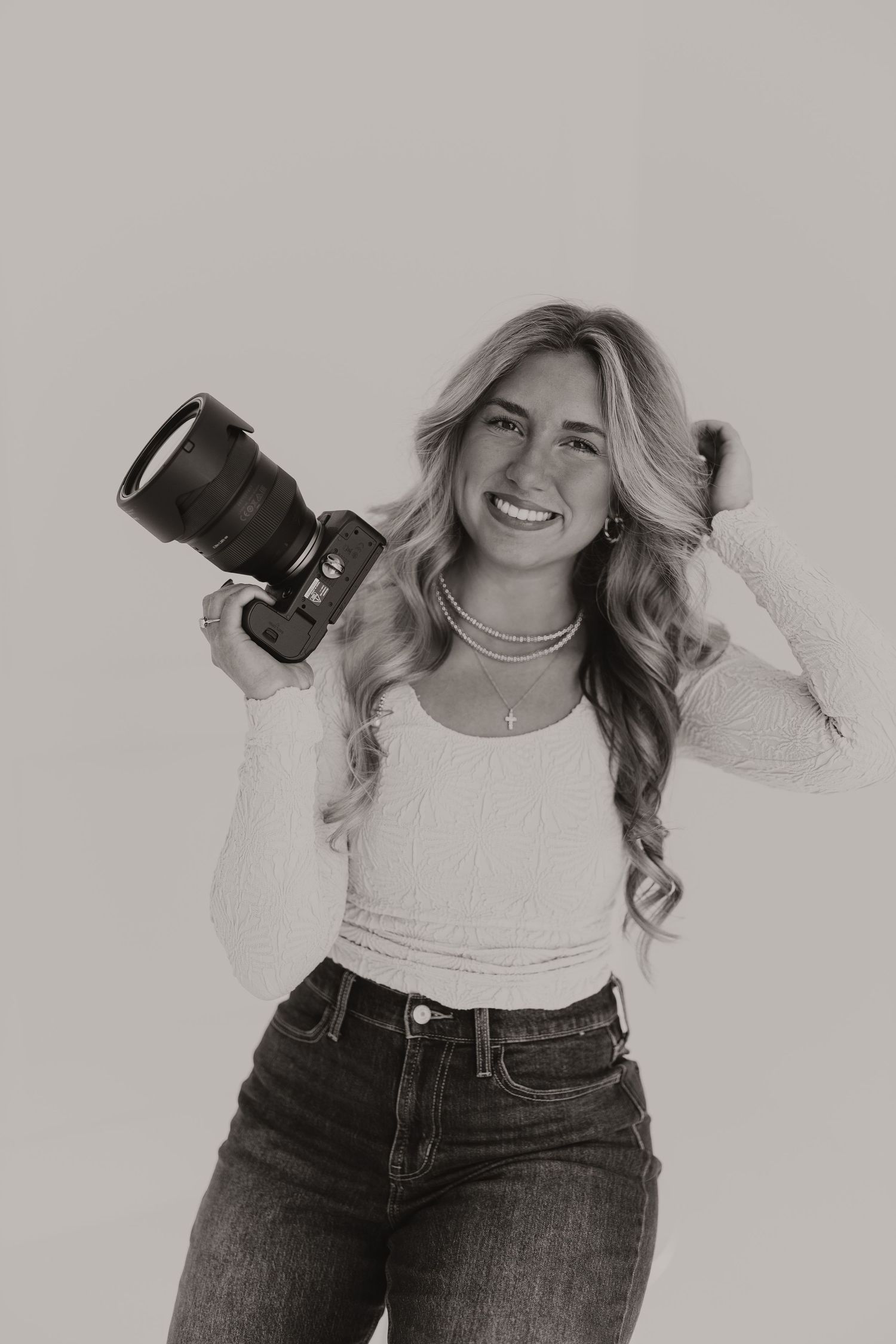 Smiling blonde woman holding DSLR camera in black and white portrait wearing white top and jeans.