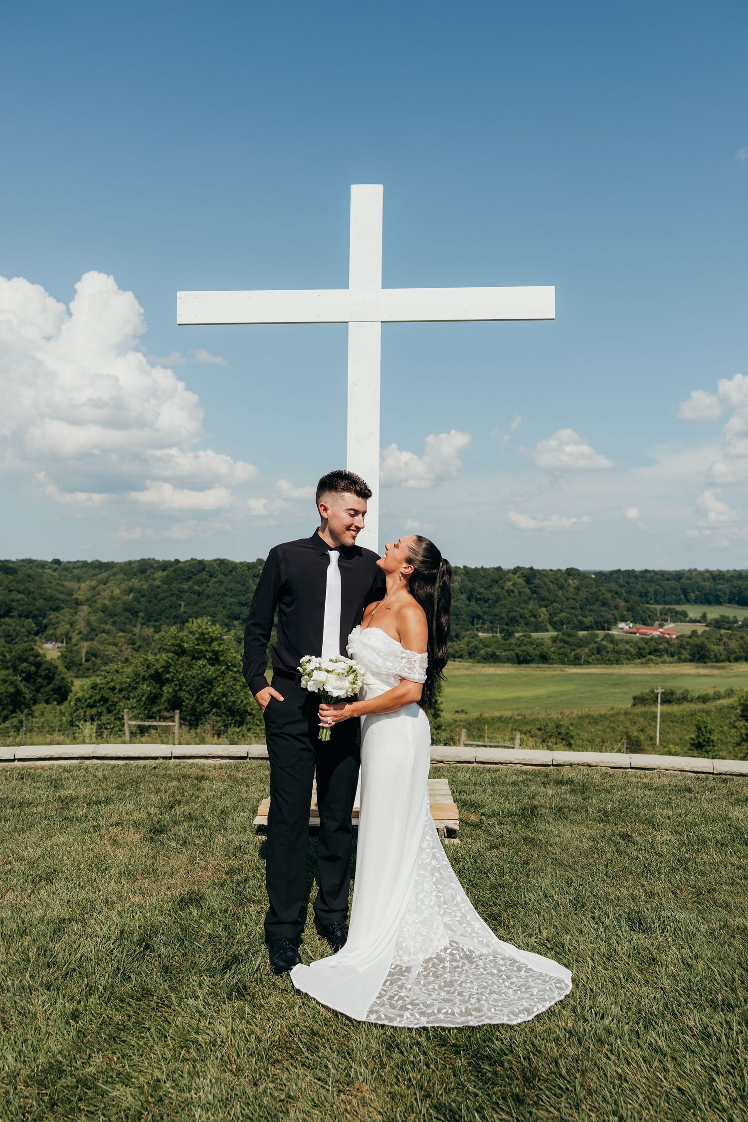 Smiling bride and groom pose together in front of a large white cross on a scenic green hilltop.