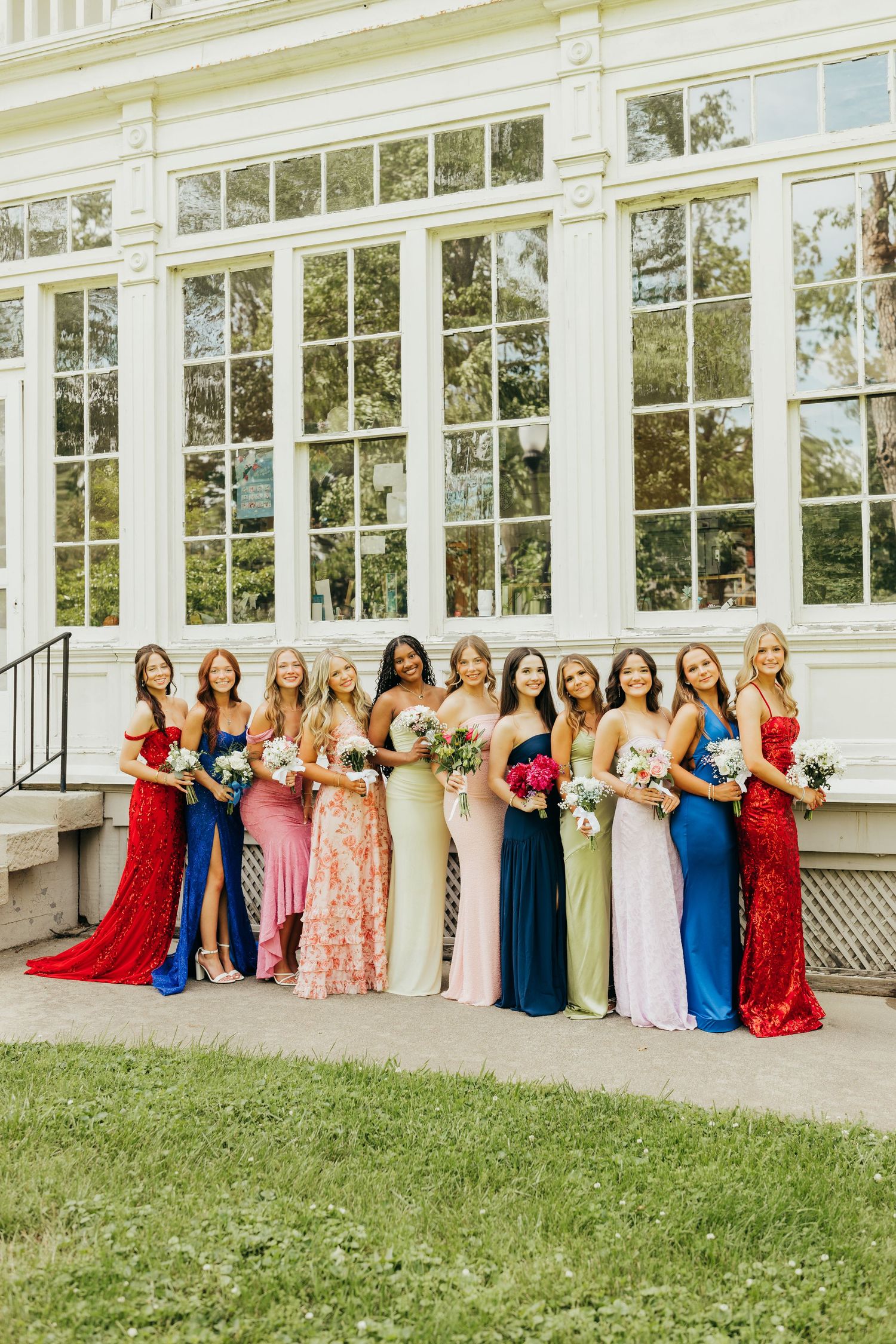 Ten bridesmaids in colorful formal gowns holding bouquets outside an elegant white building.