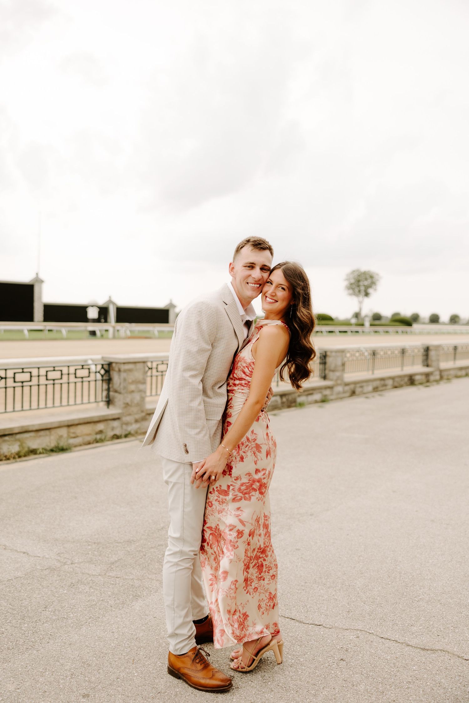 Smiling couple posing together on a wide outdoor walkway, woman in floral dress and man in beige suit.