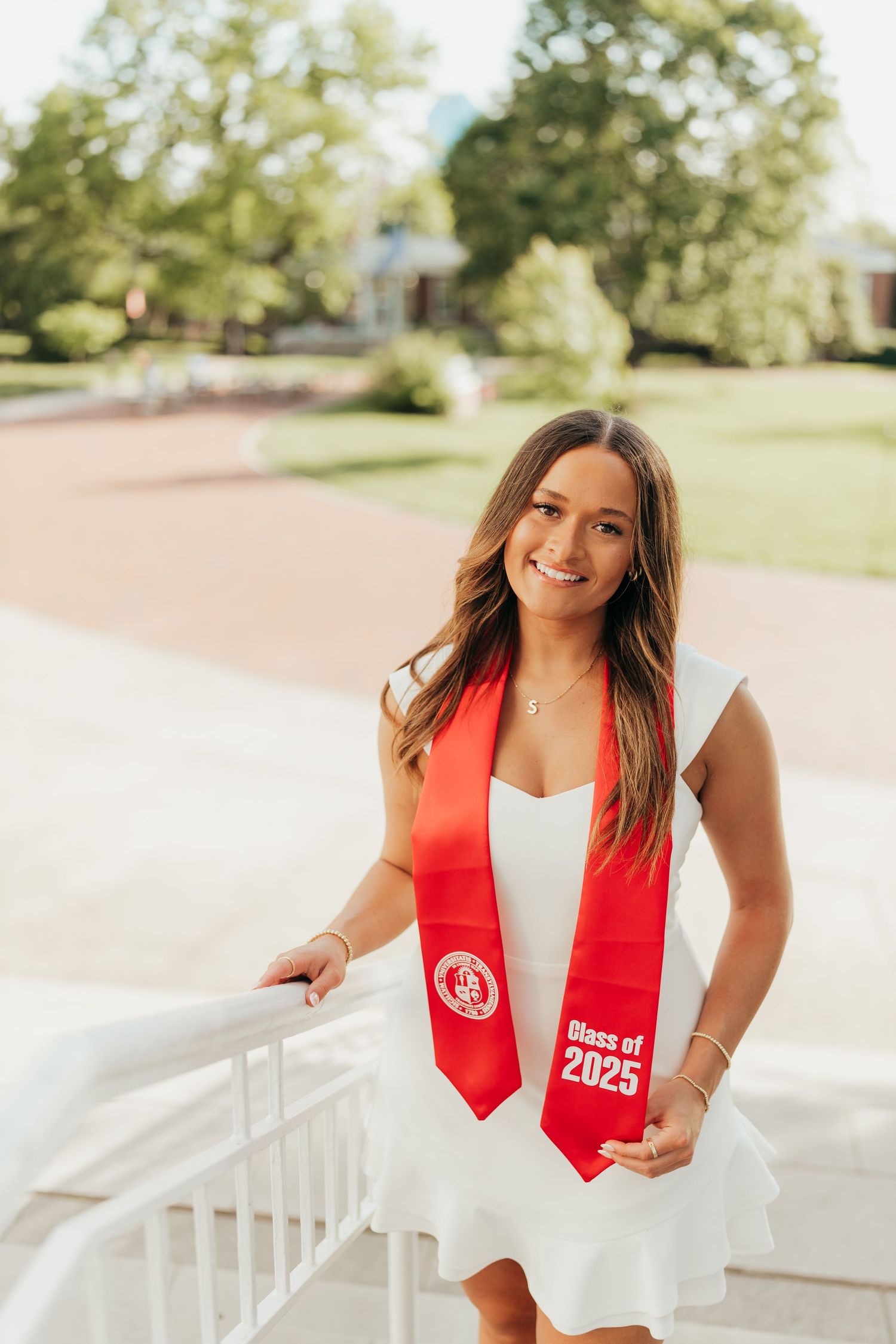 Graduate in a white dress and red graduation stole poses outdoors on campus.