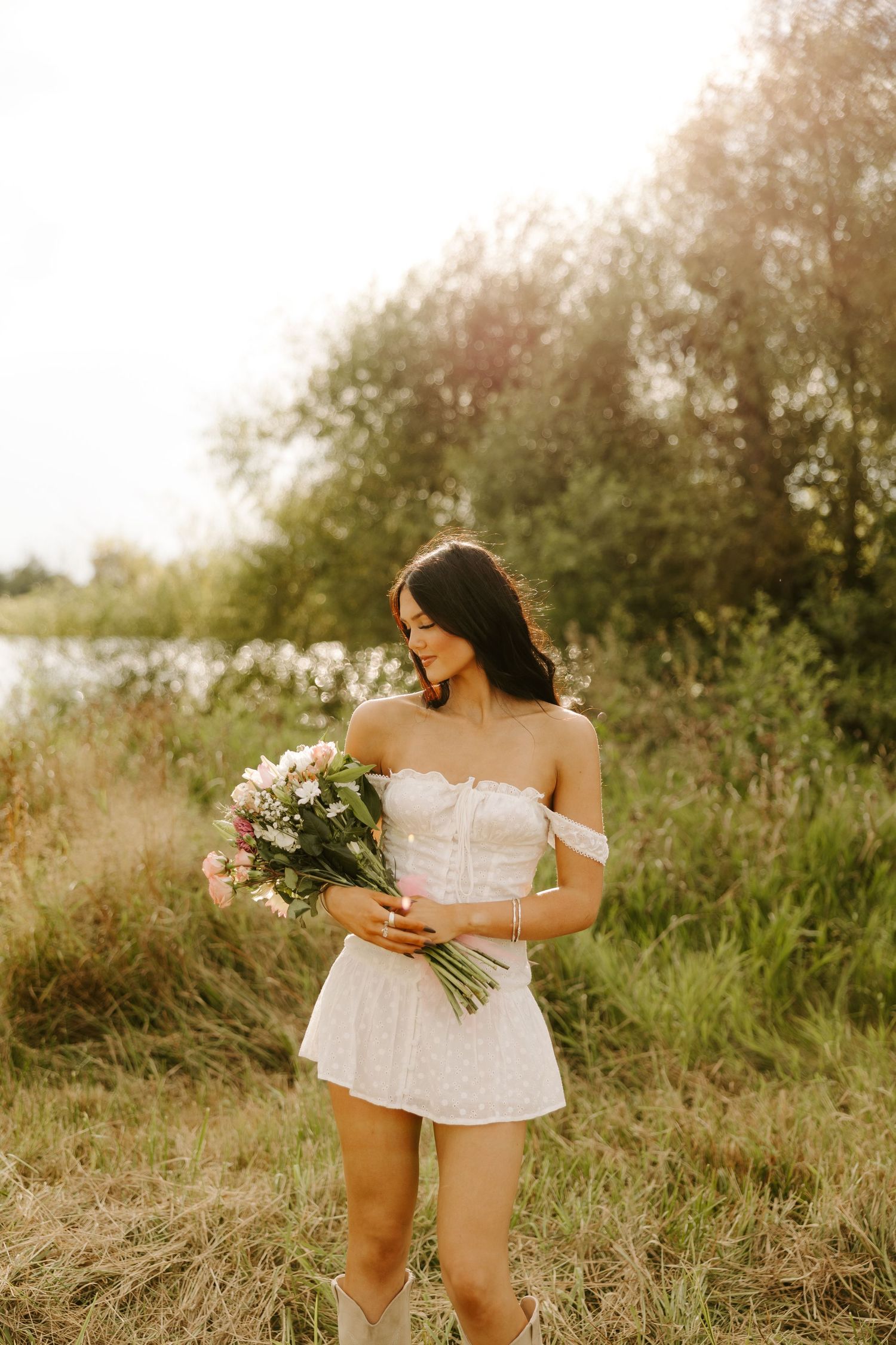 A romantic portrait in a white dress holding a fresh bouquet against a sunset meadow backdrop.