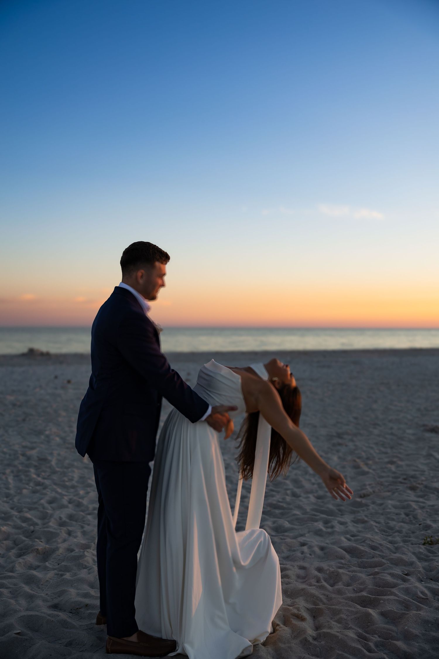 Couple dancing romantically on a sandy beach at sunset, bride in white flowing gown and groom in navy suit.