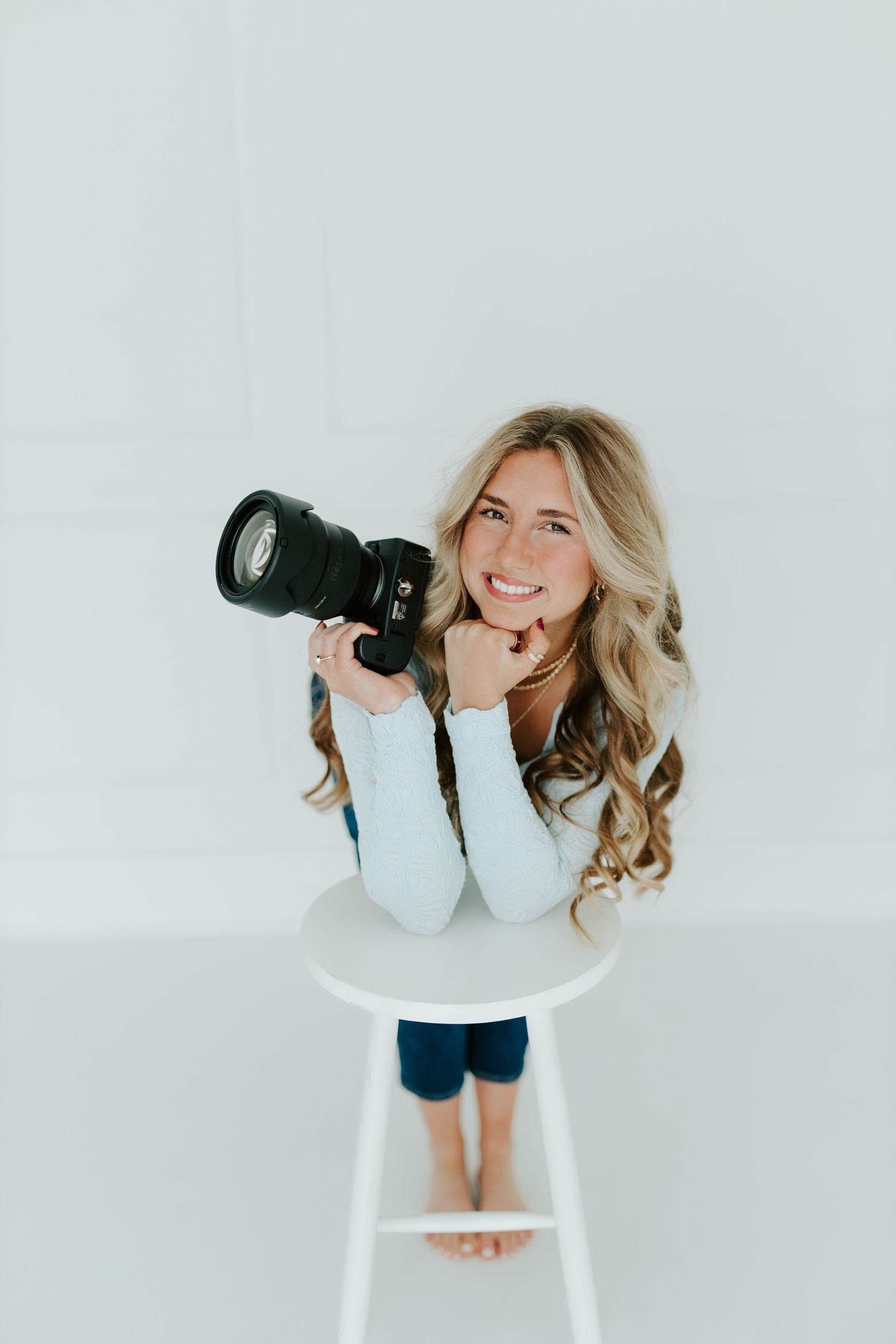 Smiling blonde woman sitting on white stool holding DSLR camera, photographed from above in studio.