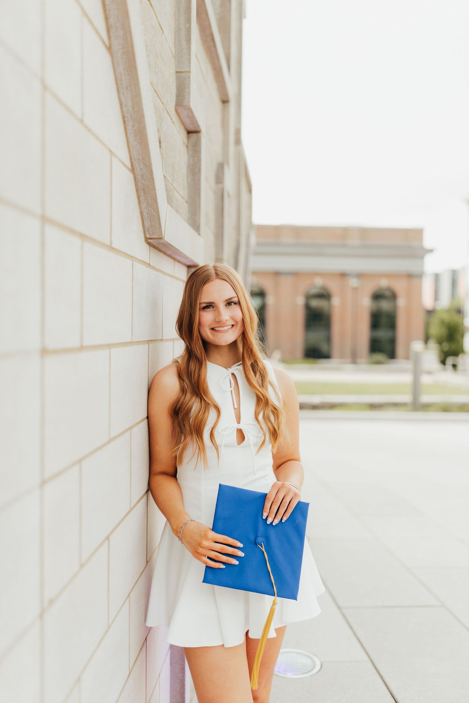 Smiling graduate in white dress holding blue graduation cap against a stone building wall.