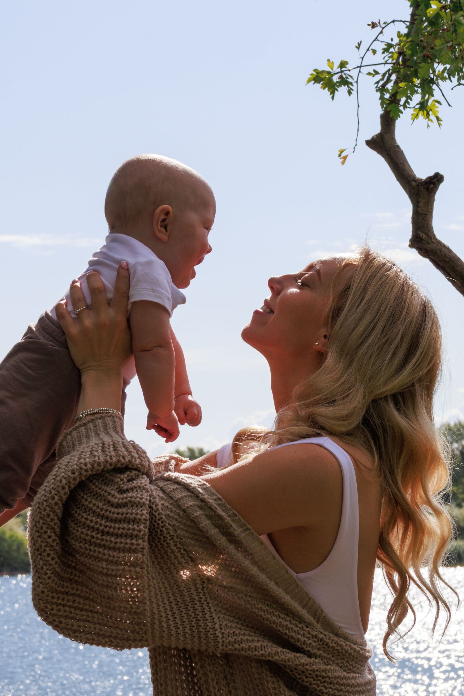 A tender moment between parent and infant by the water on a sunny day with trees in the background.