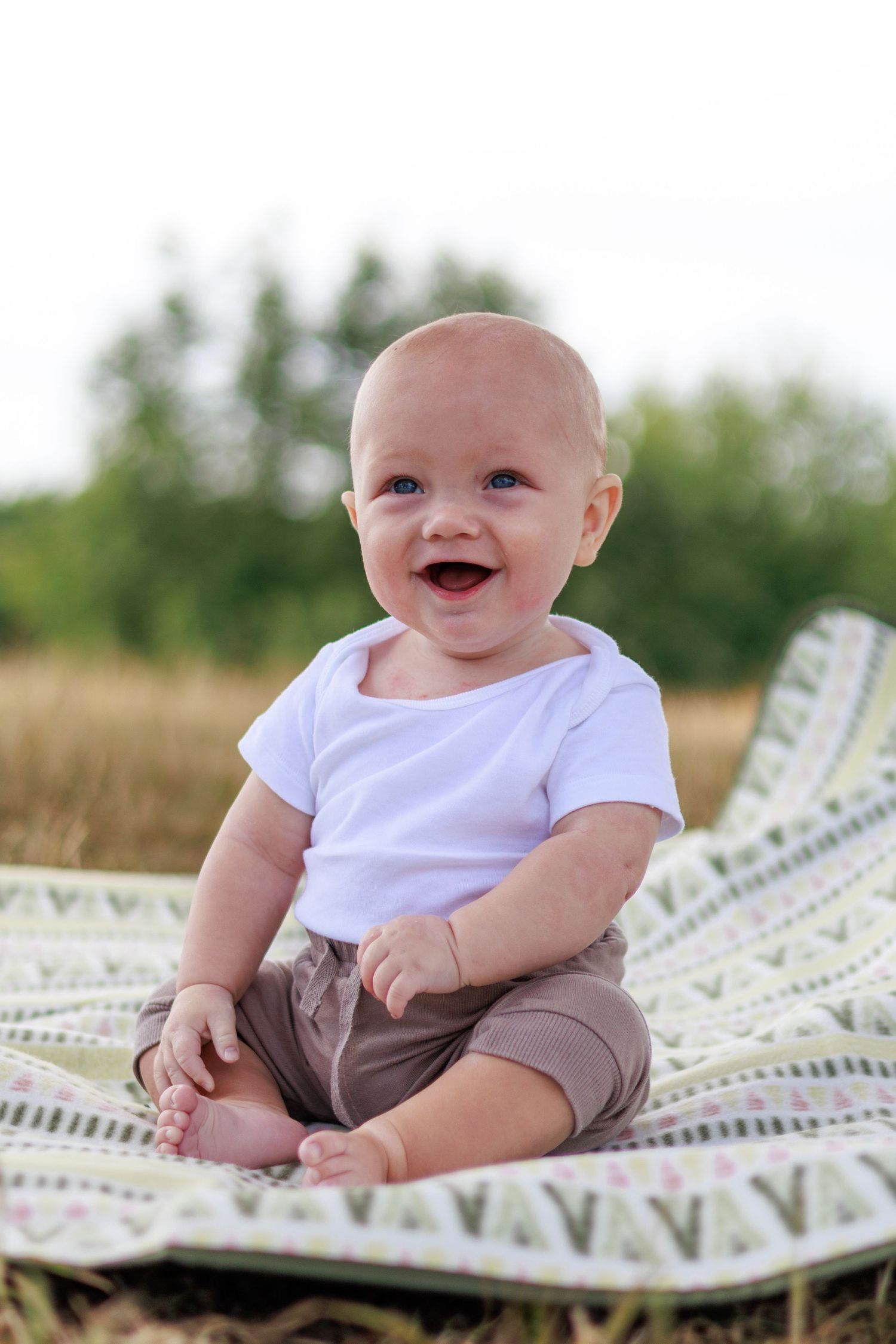 A baby in a white shirt and gray pants lies on a patterned blanket outdoors with a joyful expression.