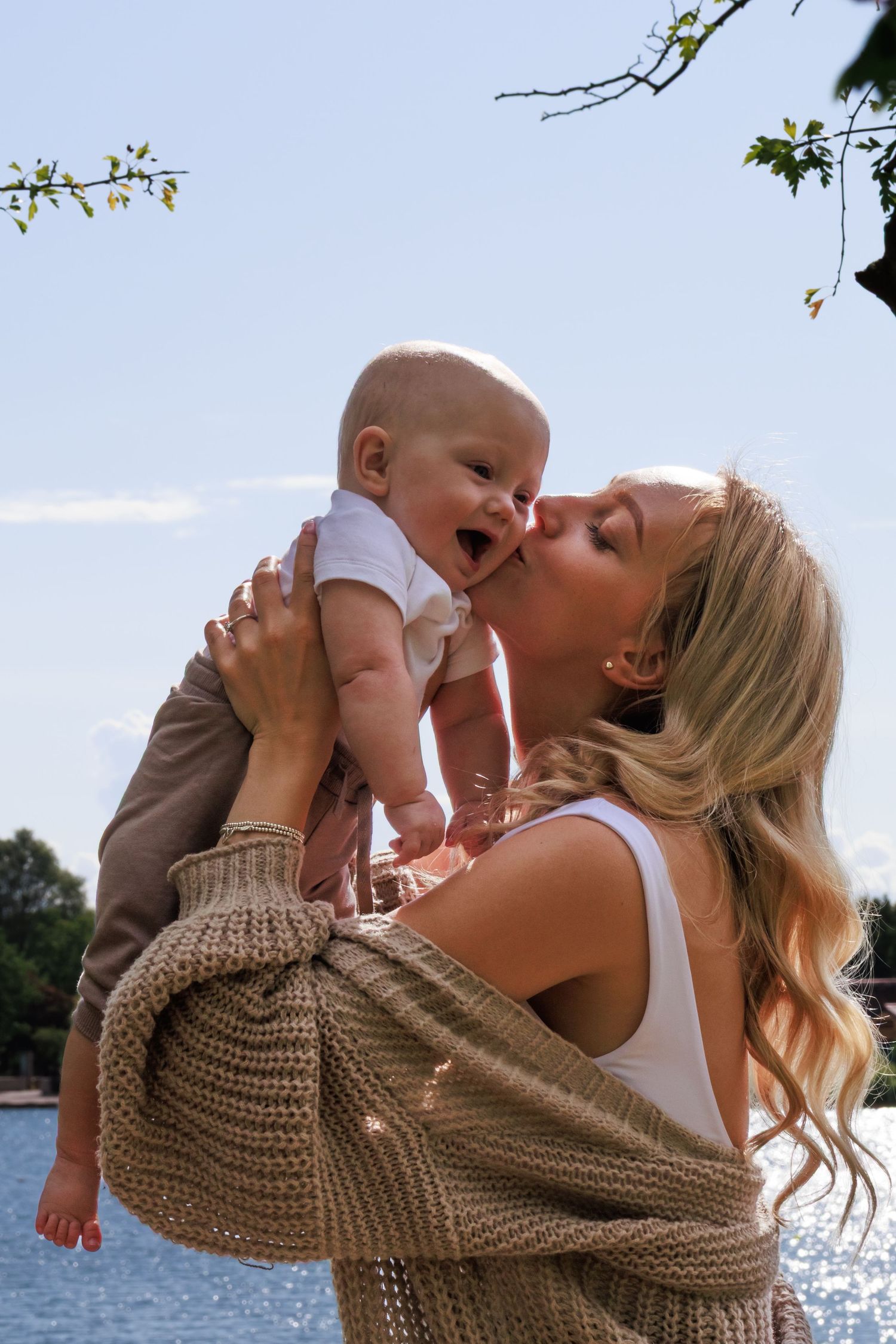 A tender moment of joy and laughter between parent and baby by a sparkling waterfront on a sunny day.