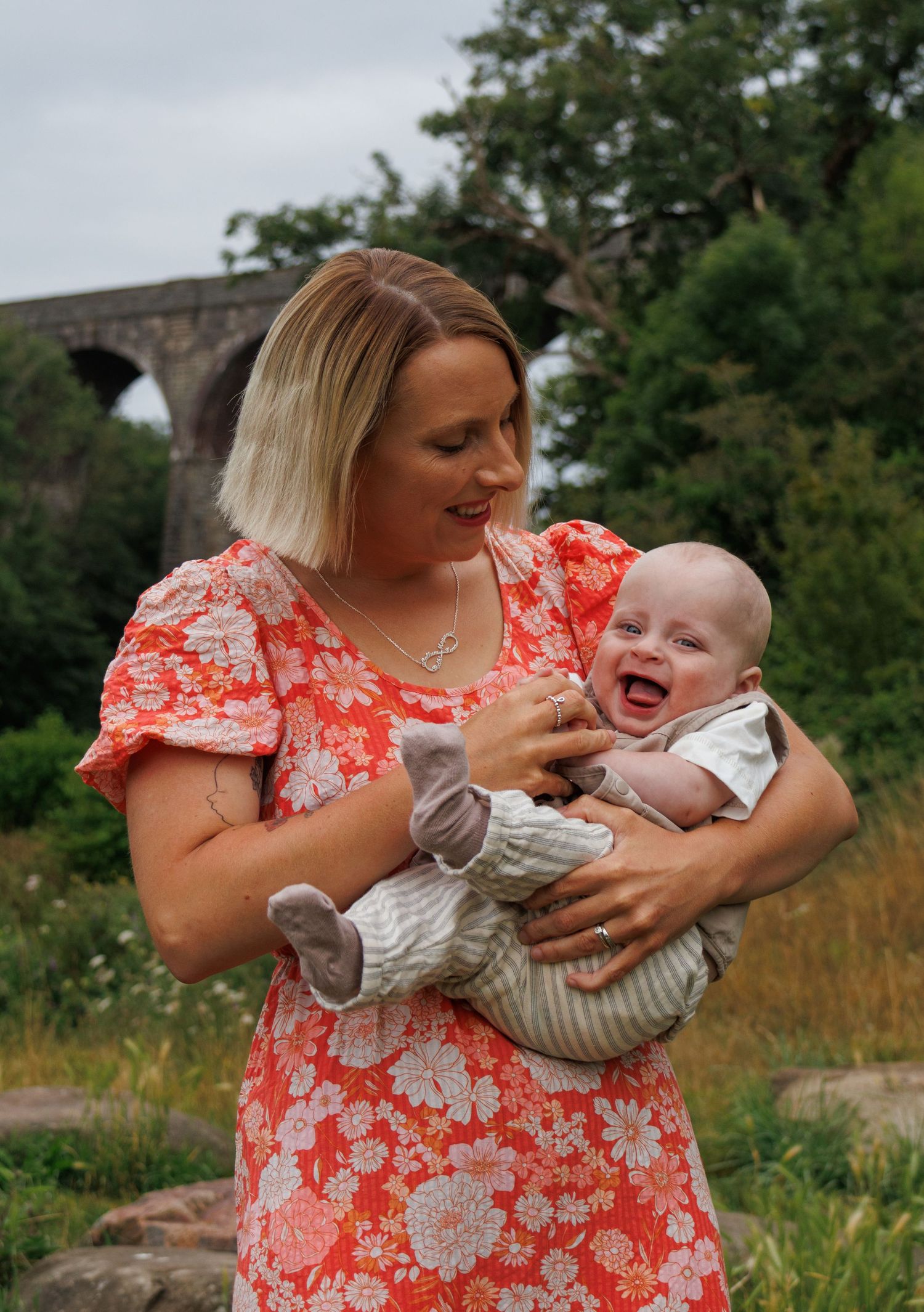A mother in a red floral dress playfully holds and tickles a laughing baby in a striped outfit outdoors.