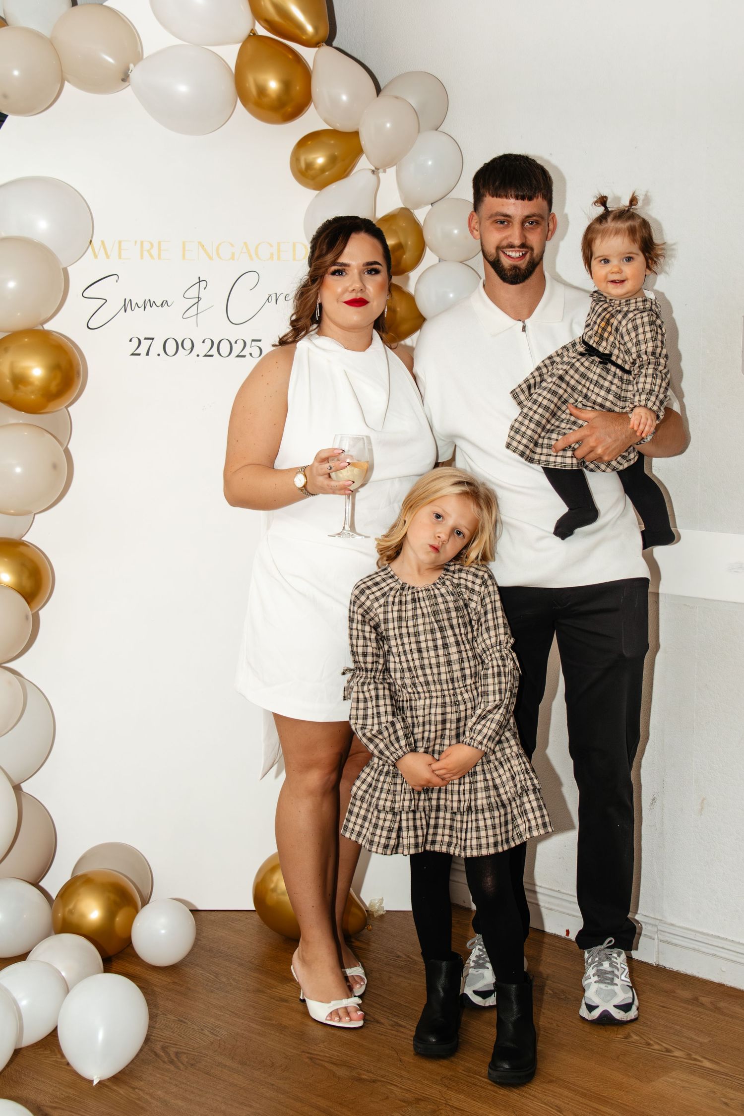 Elegant family photo setup with white and gold balloon arch decorations and matching gingham dresses for a special occasion.