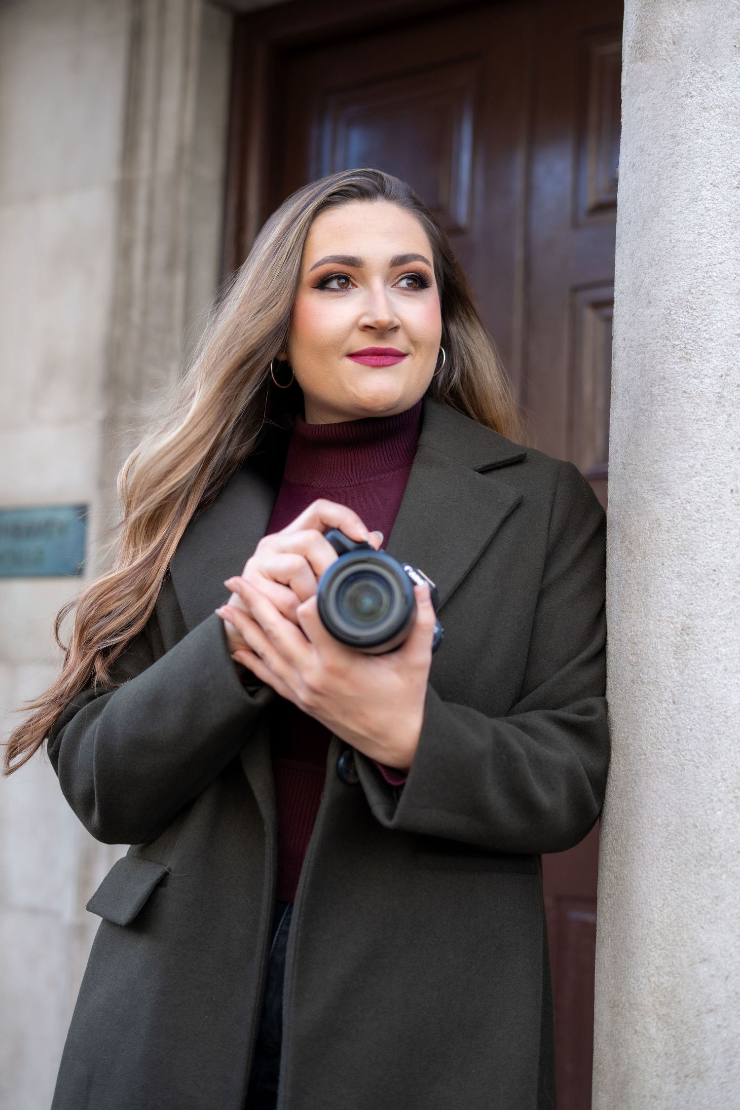 Four sequential photos of someone in a black coat holding a camera and posing against a light colored wall.