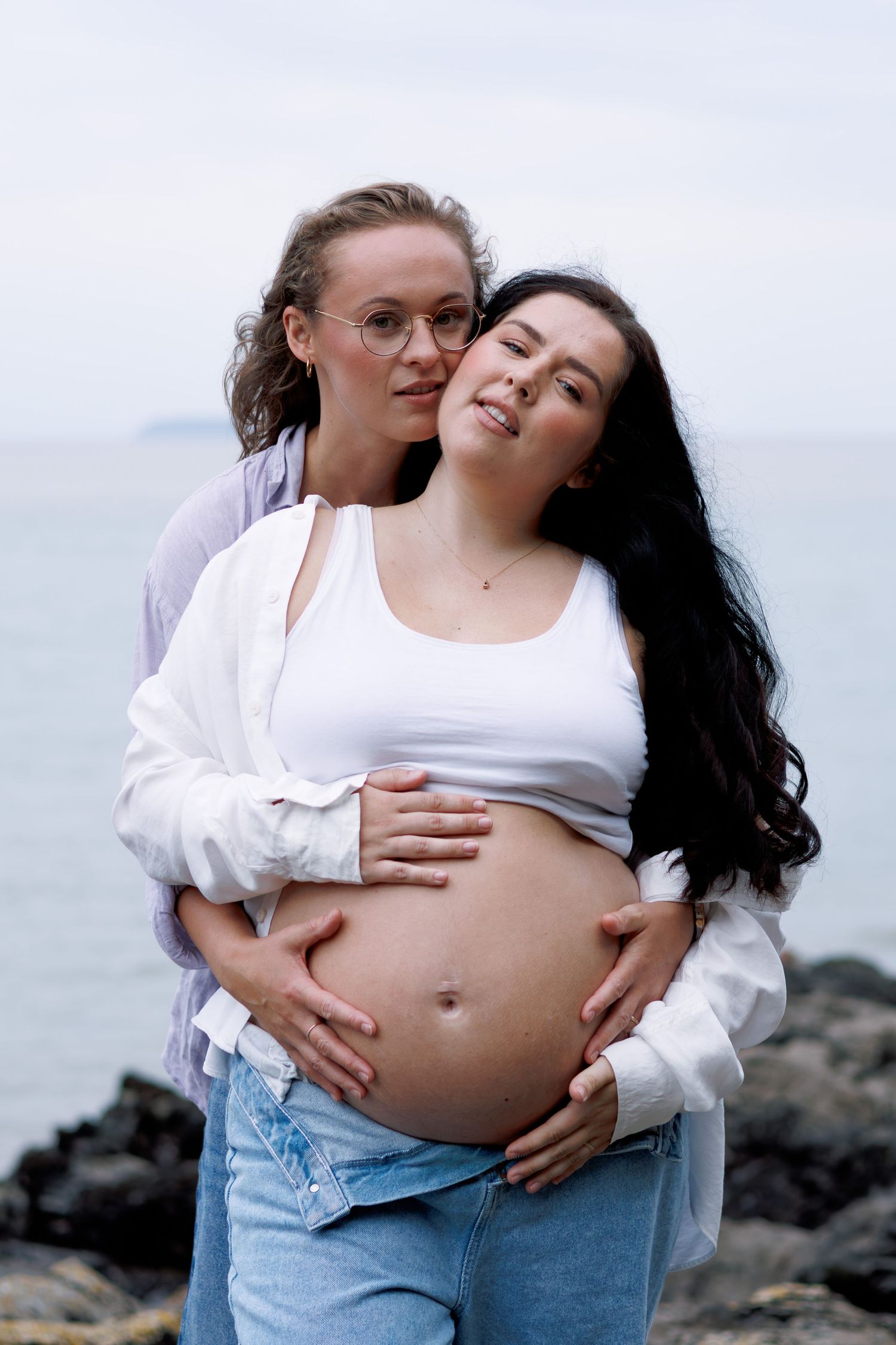A loving maternity photo taken on rocky beach shore with overcast sky in the background.