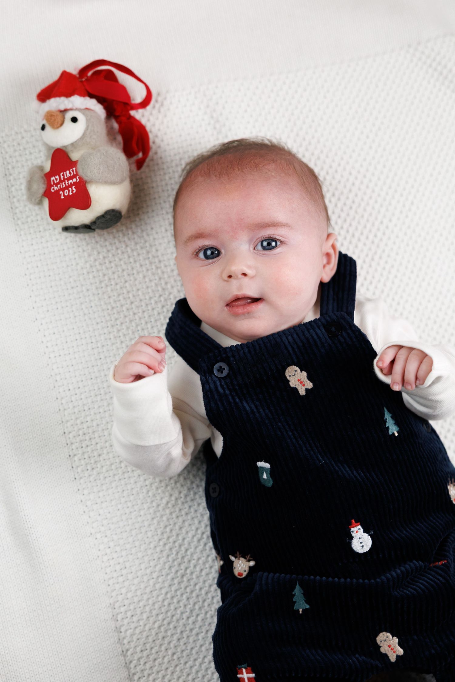 A baby in a navy sweater vest with holiday motifs lies on white bedding next to a small plush snowman ornament.