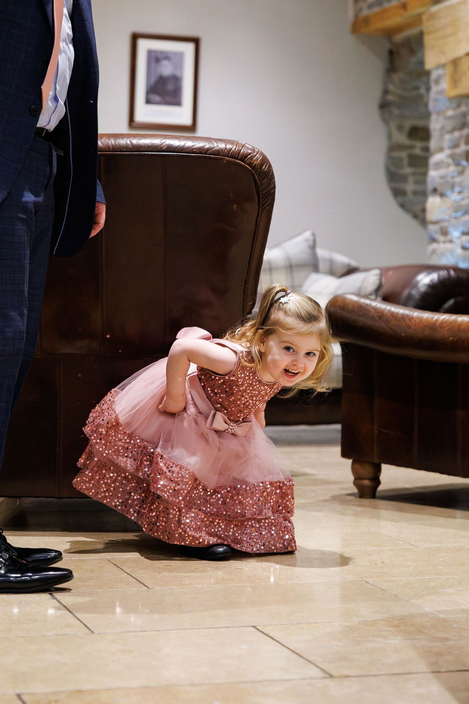 A young girl in a pink lace dress lies playfully on a stone floor while someone in dark pants stands nearby.