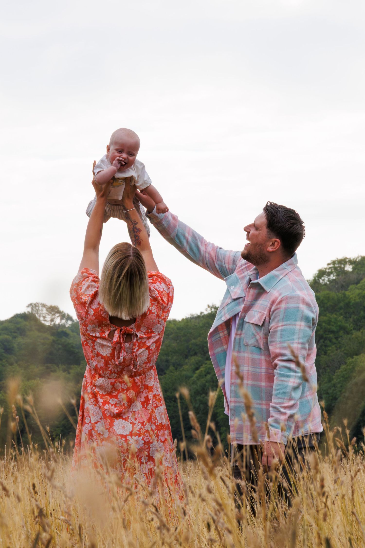 A couple lifts their baby together in a field of tall grass during sunset, wearing colorful floral and plaid outfits.