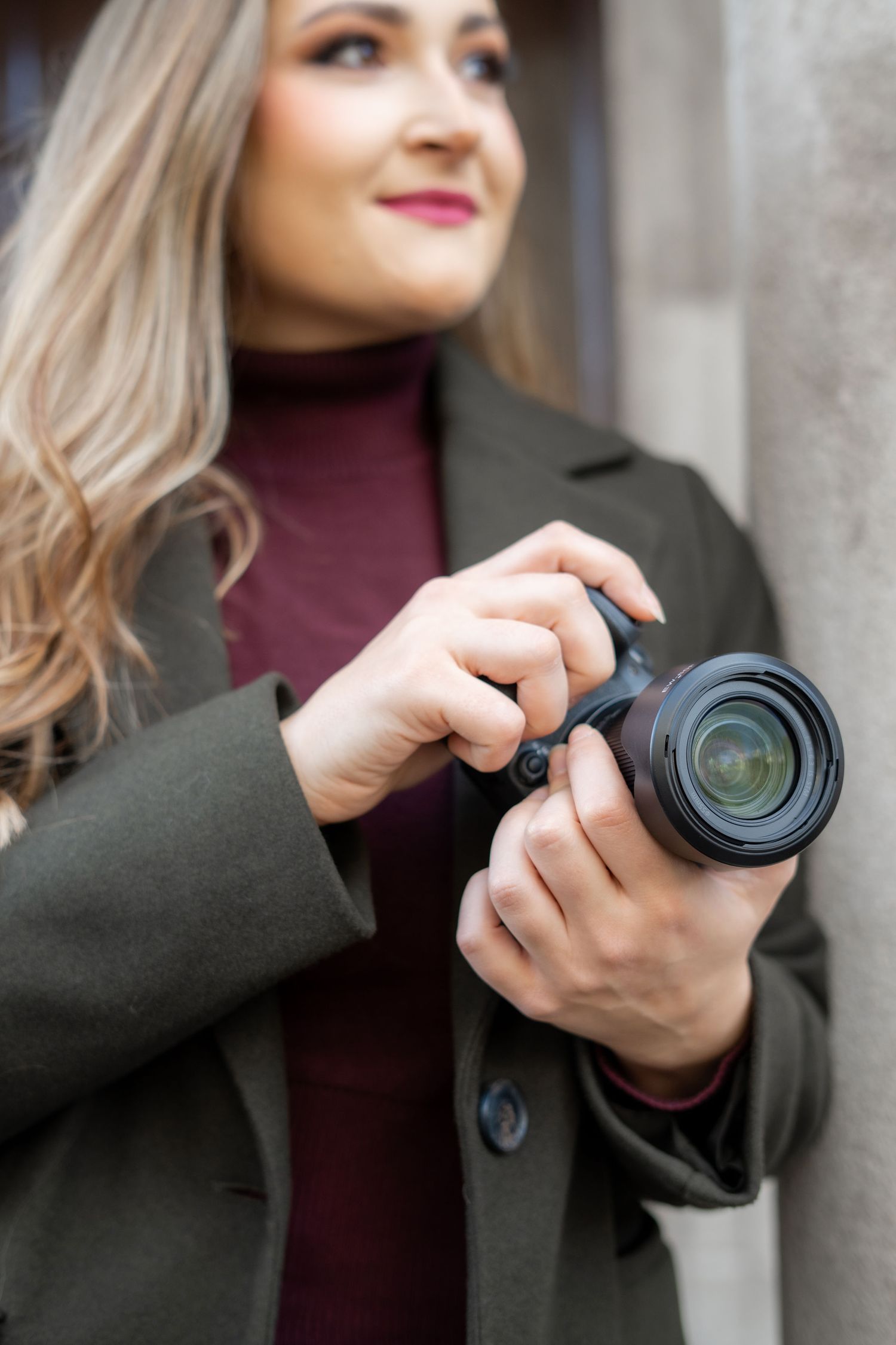 Professional photographer in burgundy turtleneck and olive coat adjusts camera settings while shooting outdoors.