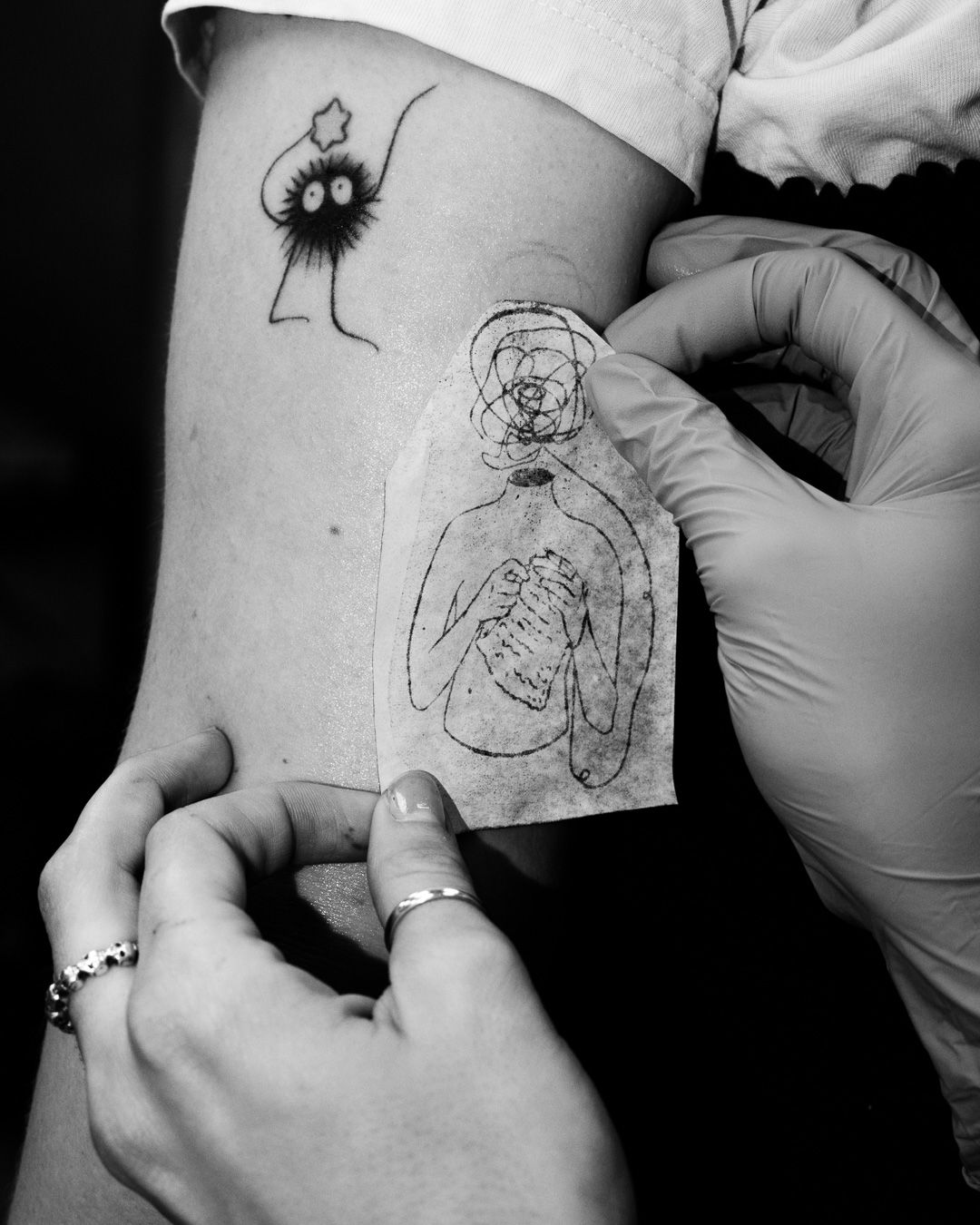 Black and white photo of a tattoo artist working on a delicate line drawing tattoo with gloved hands.