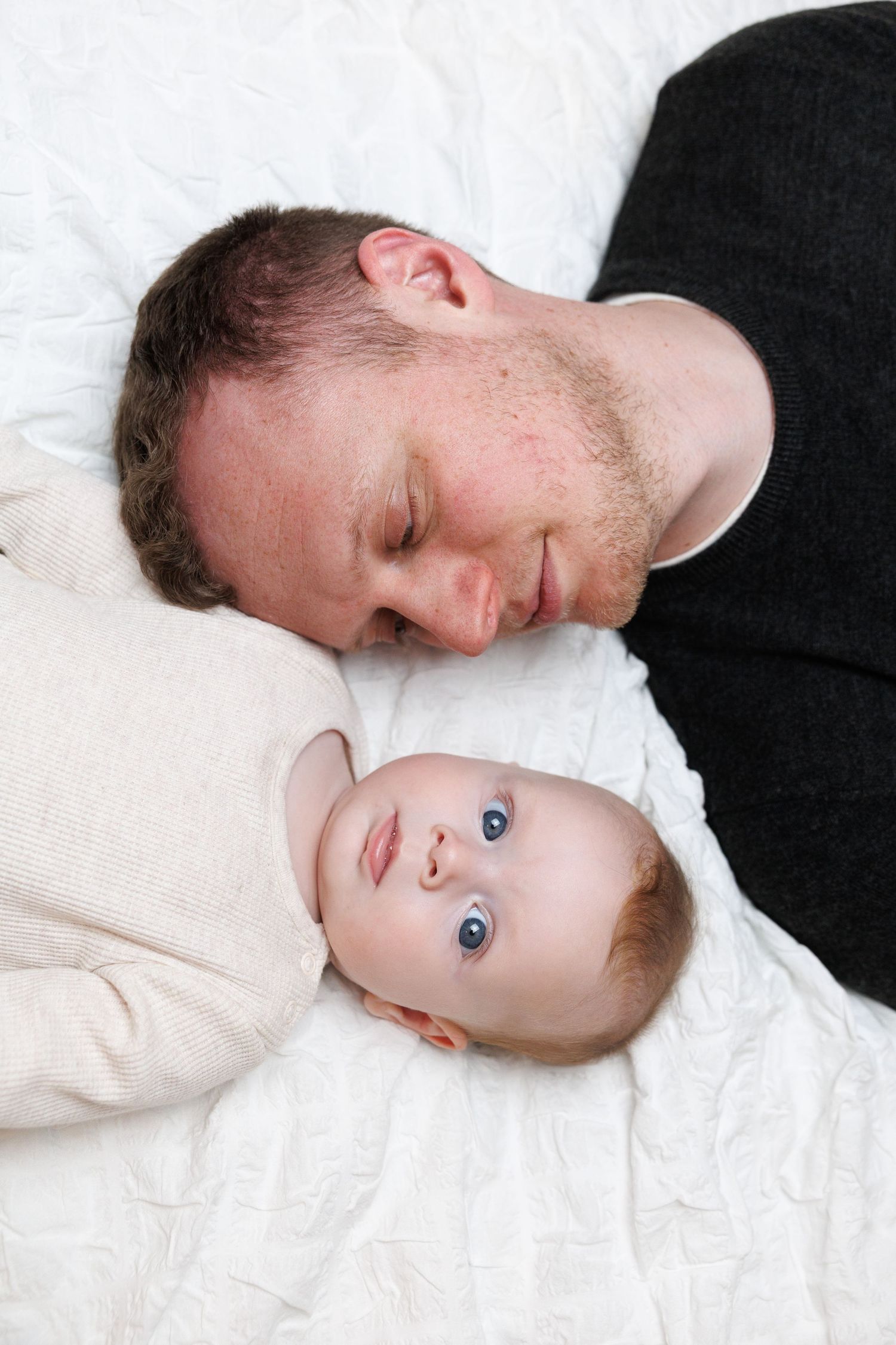 A tender moment between an adult and baby lying together on white bedding.