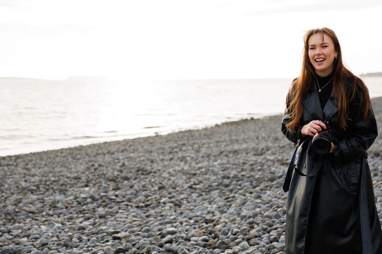 A person in a black coat walks along a rocky beach on a cloudy day.