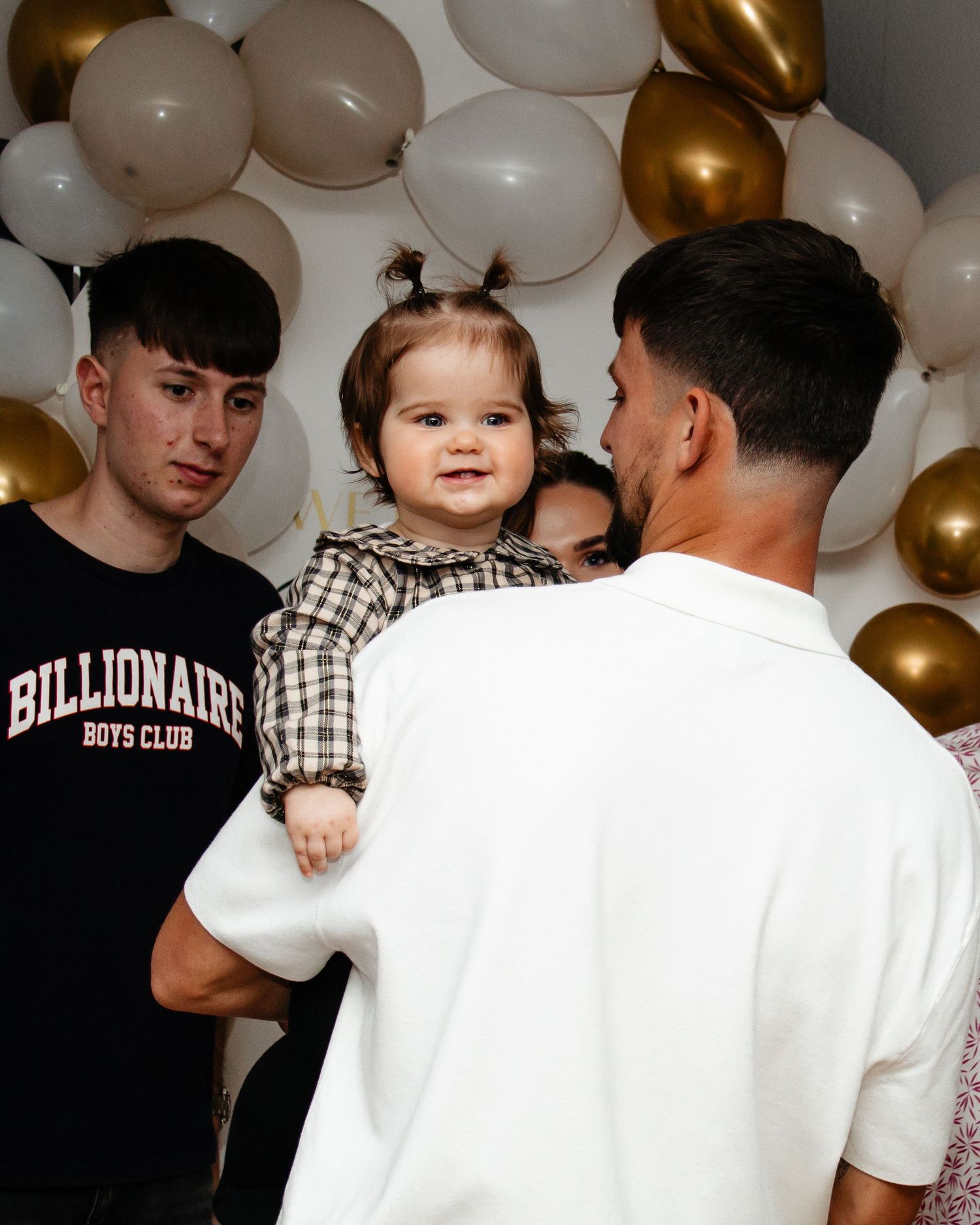 A party scene with gold and white balloons decorating the background as people gather together.