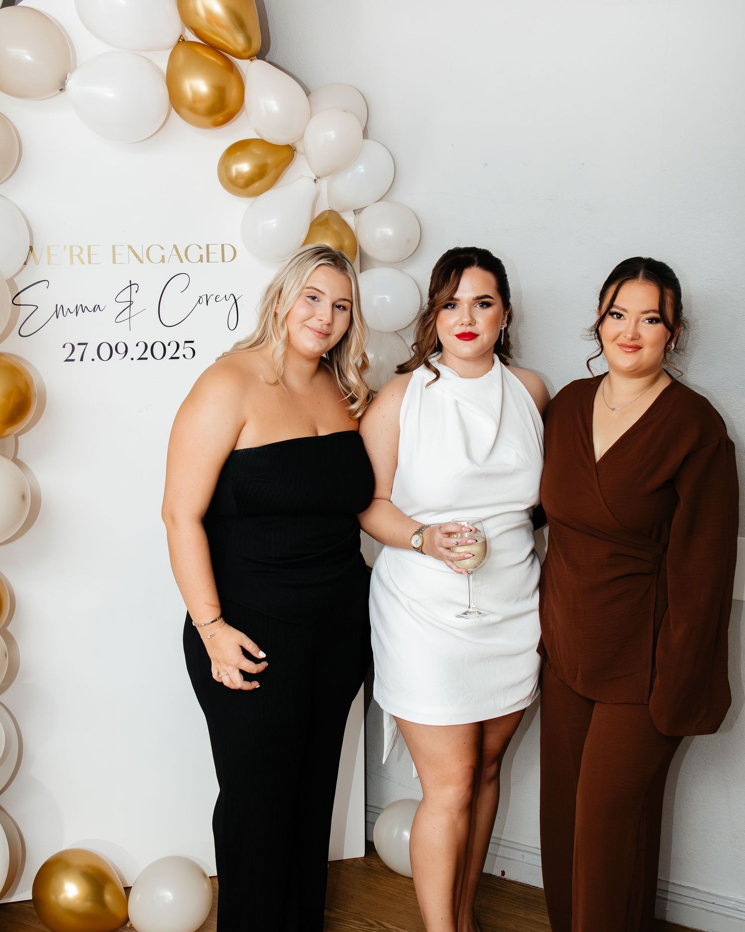 Three people pose together against a white wall decorated with gold and white balloons for an engagement celebration.