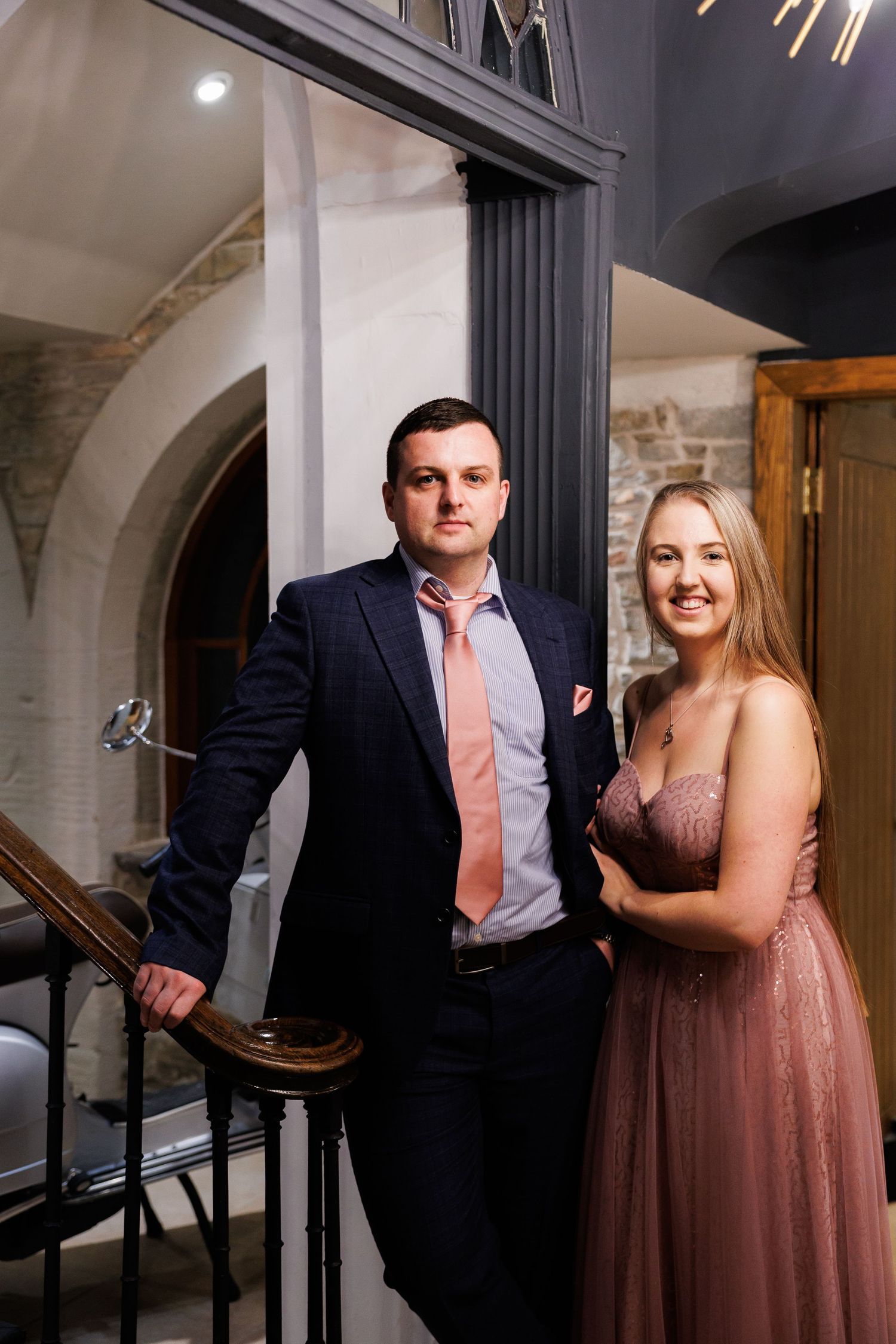Elegant couple poses on staircase at formal event, with woman in dusty rose gown and man in dark suit.
