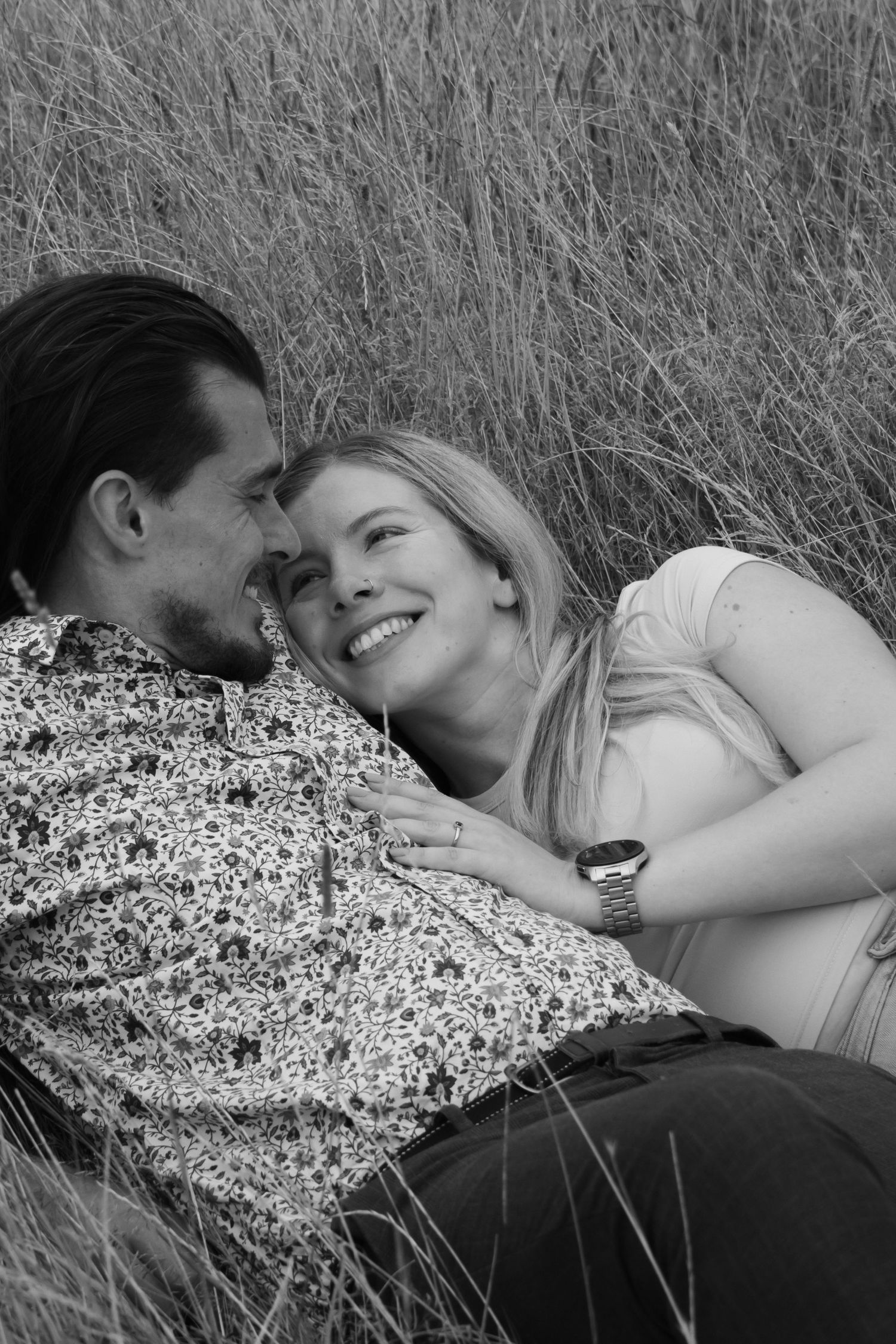 A romantic black and white photo of a couple lying in tall grass together, sharing an intimate moment.