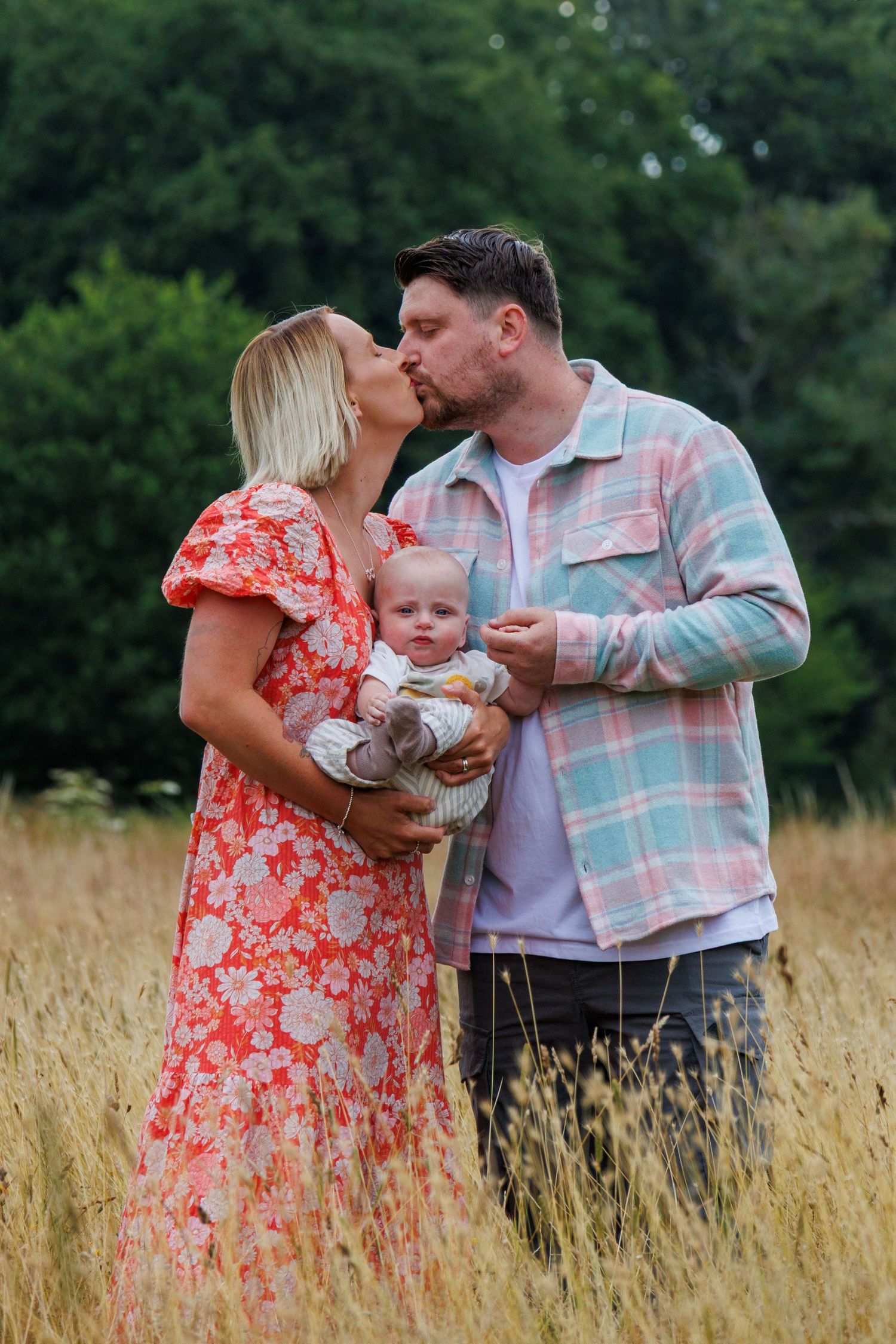 A couple and their baby share tender moments in a golden wheat field while wearing coral and plaid outfits.