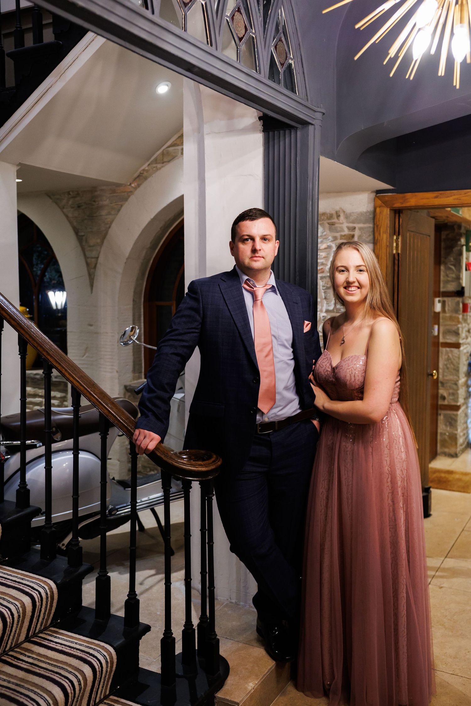 A couple in formal attire poses together in front of a staircase in an elegant indoor setting.