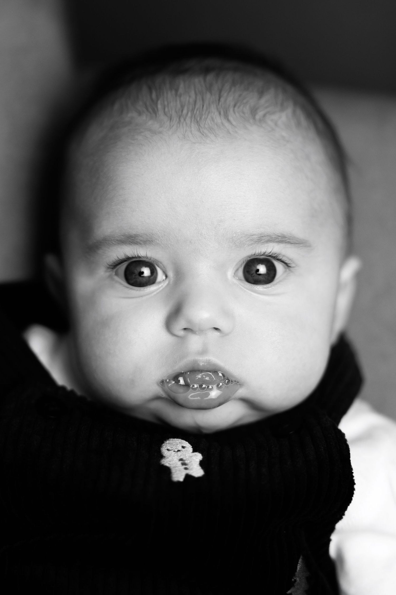 A black and white close-up portrait of a baby with wide eyes and pursed lips wearing a dark sweater.