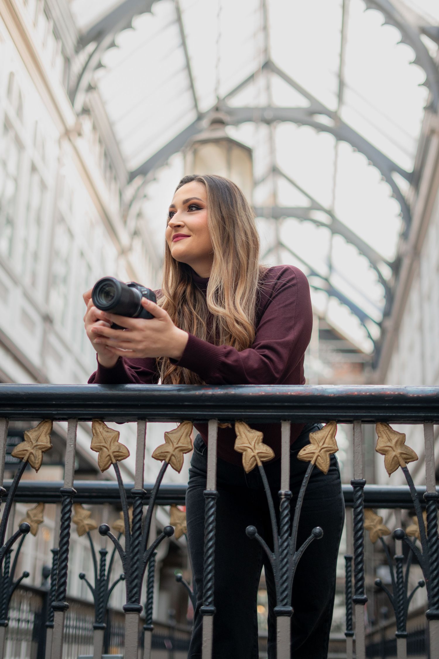 A photographer in a burgundy sweater leans on a metal railing while taking photos in an architectural setting.