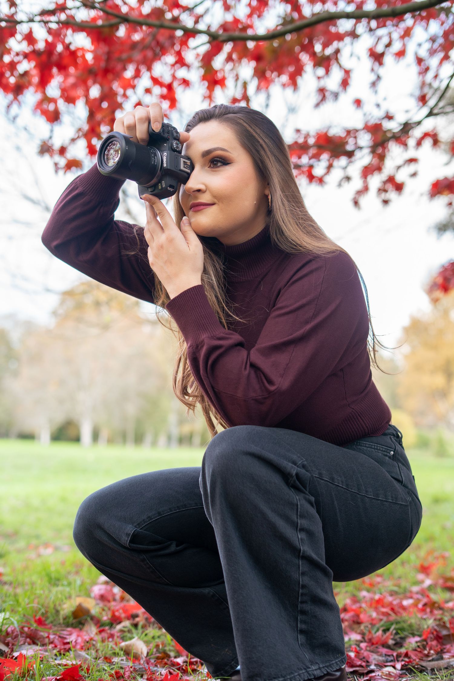 A photographer in a burgundy sweater crouches low to capture autumn scenes among red maple leaves.